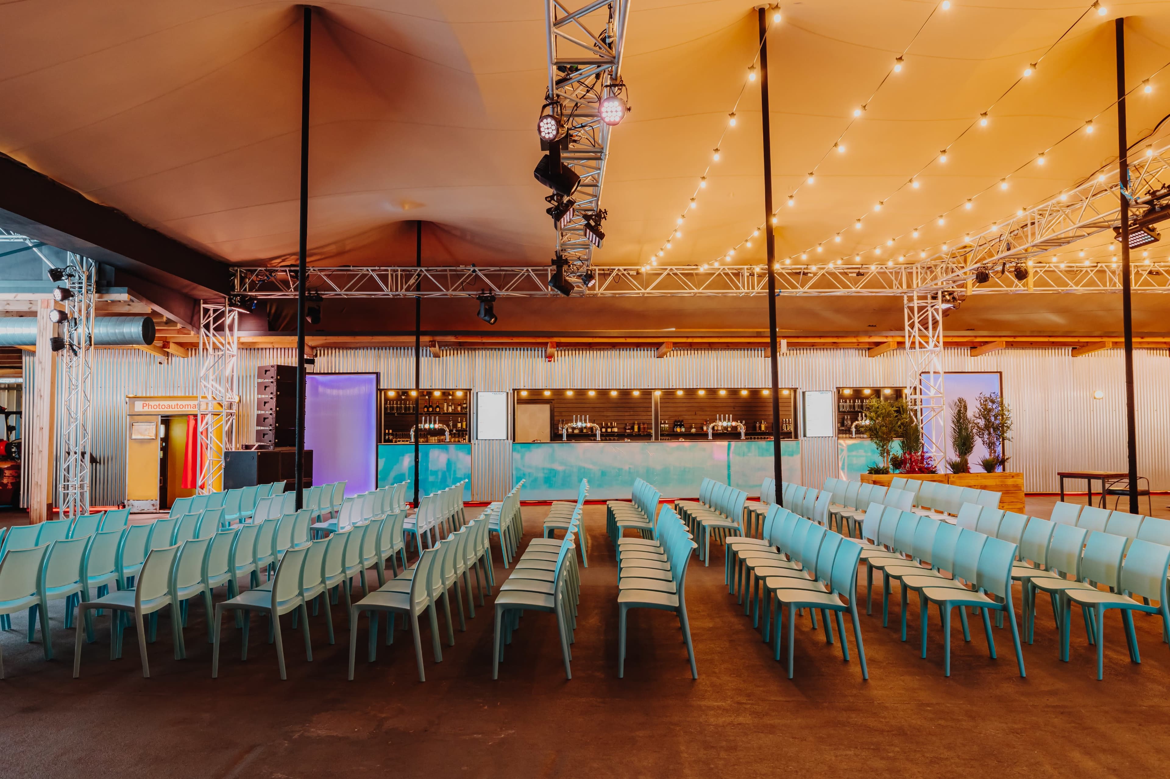 The image shows a spacious indoor venue with rows of light blue chairs arranged facing a stage area, illuminated by overhead lights and surrounded by a modern design.