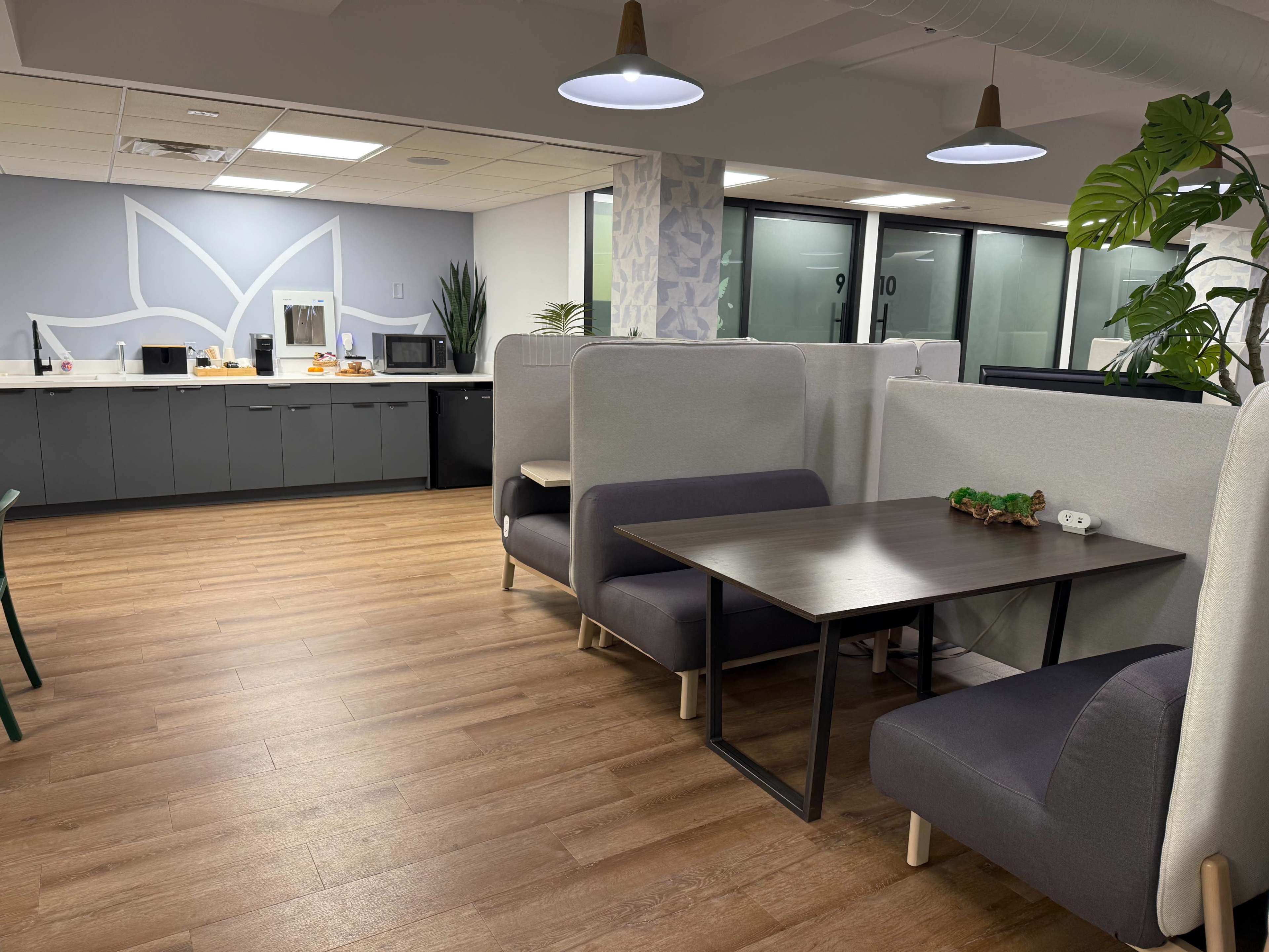 The image shows a modern office kitchen area with gray cabinetry, a microwave, and seating booths arranged around a table on a hardwood floor.