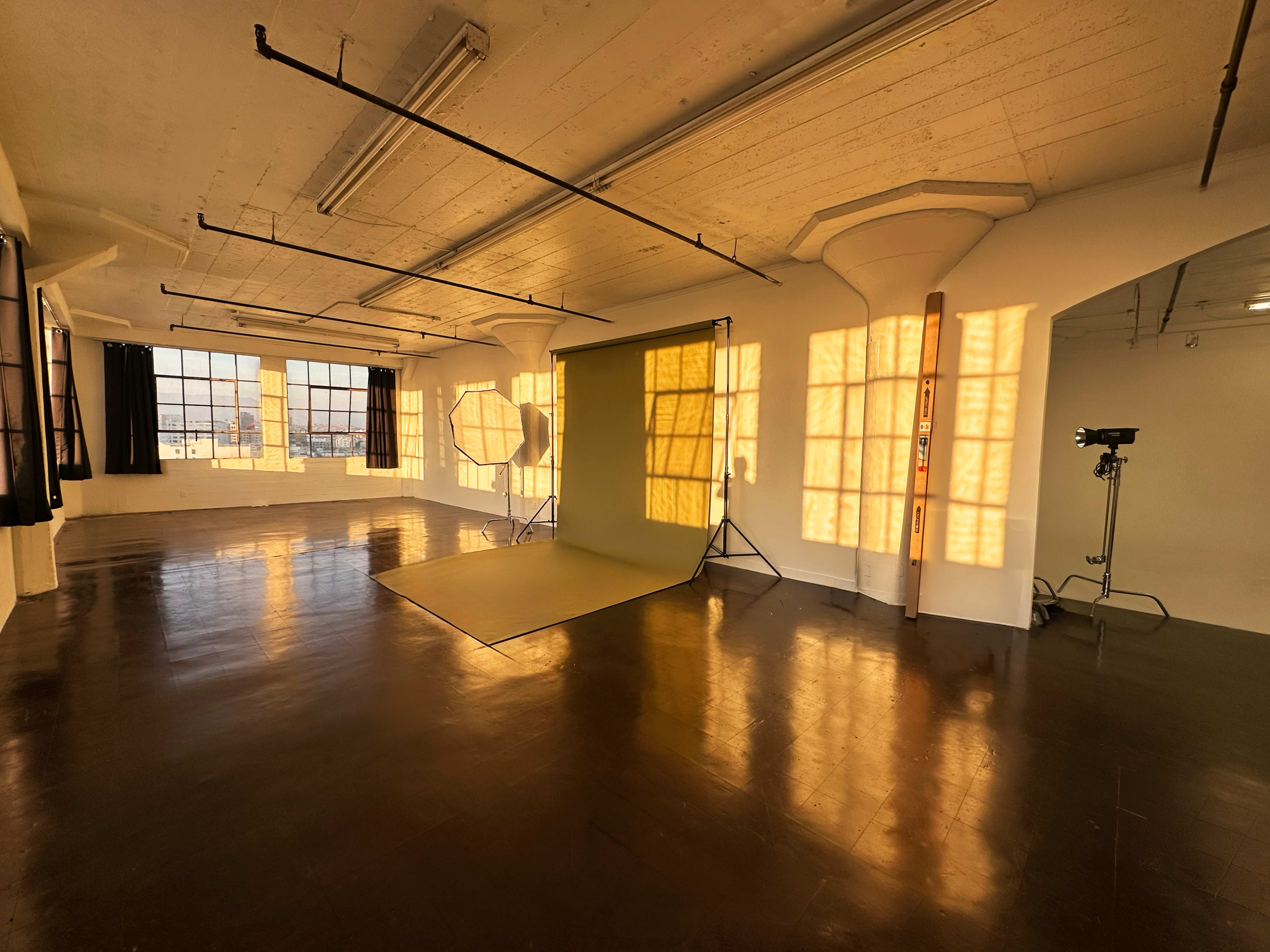 The image shows an empty studio space with large windows, a green backdrop set up, and sunlight casting geometric shadows on the floor.