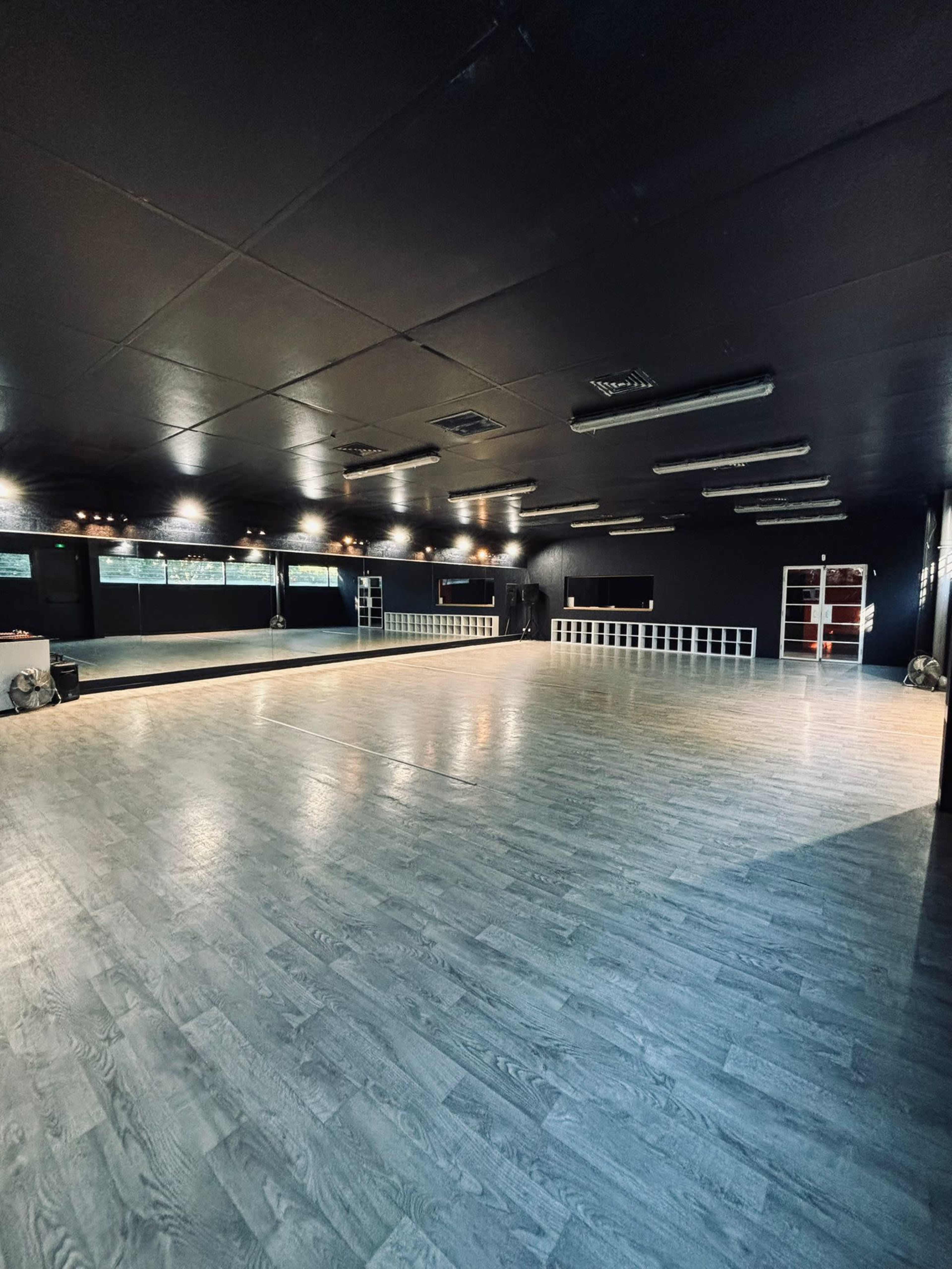 The image shows a spacious dance studio with dark walls, wooden flooring, and large mirrors along one side.