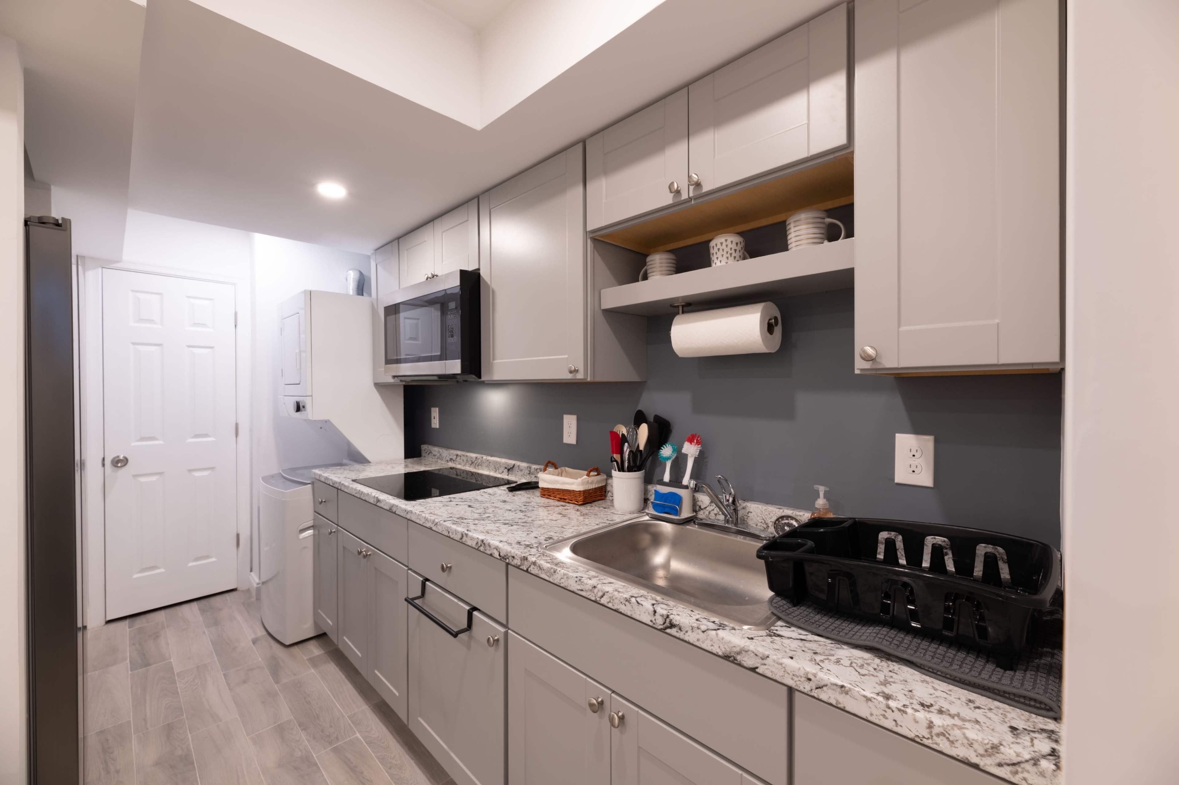 The image shows a modern kitchen with gray cabinets, a granite countertop, a sink, and appliances including a microwave and a laundry area.
