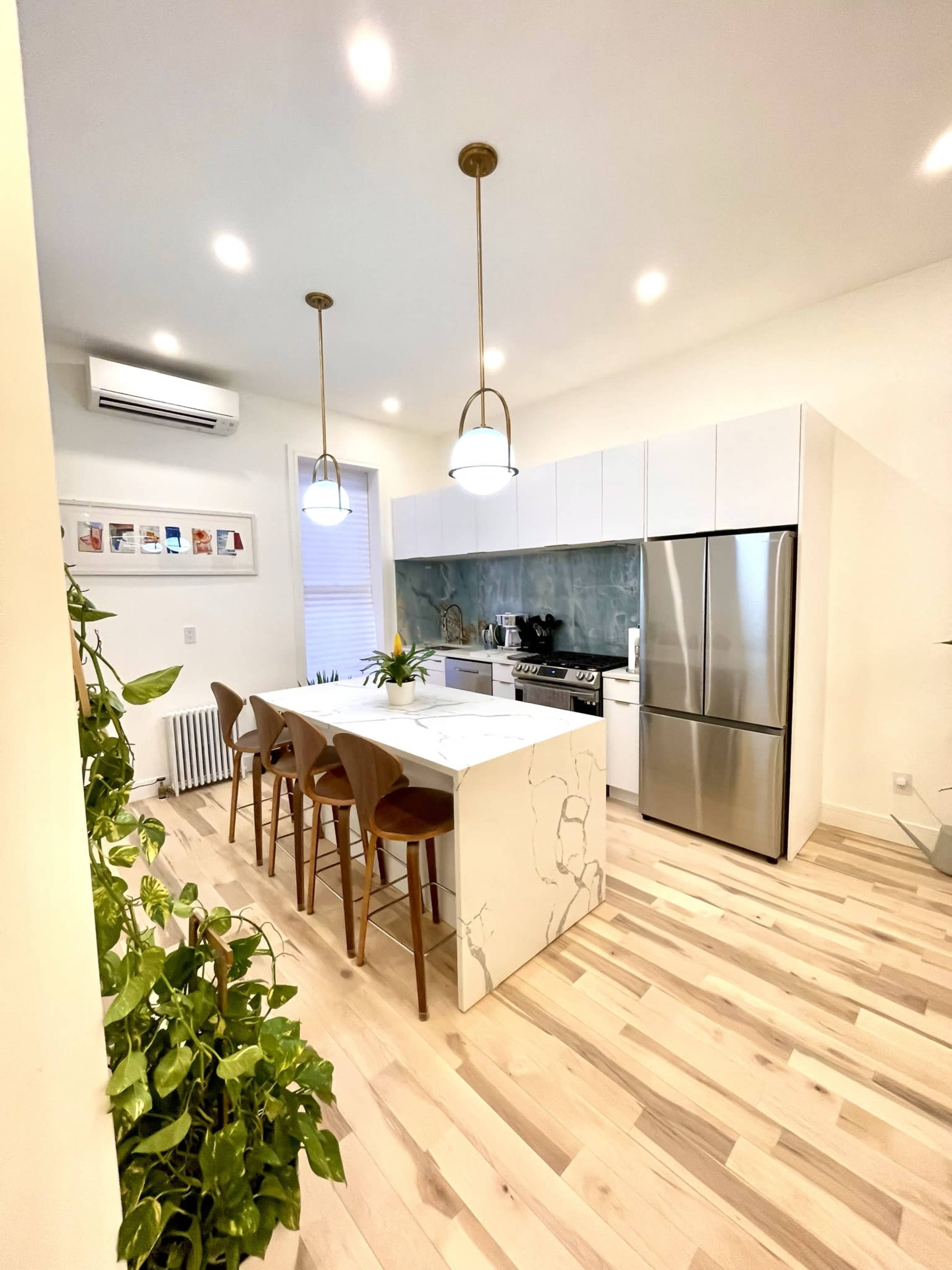A modern kitchen features a central island with bar stools, stainless steel appliances, and green plants along the wall.