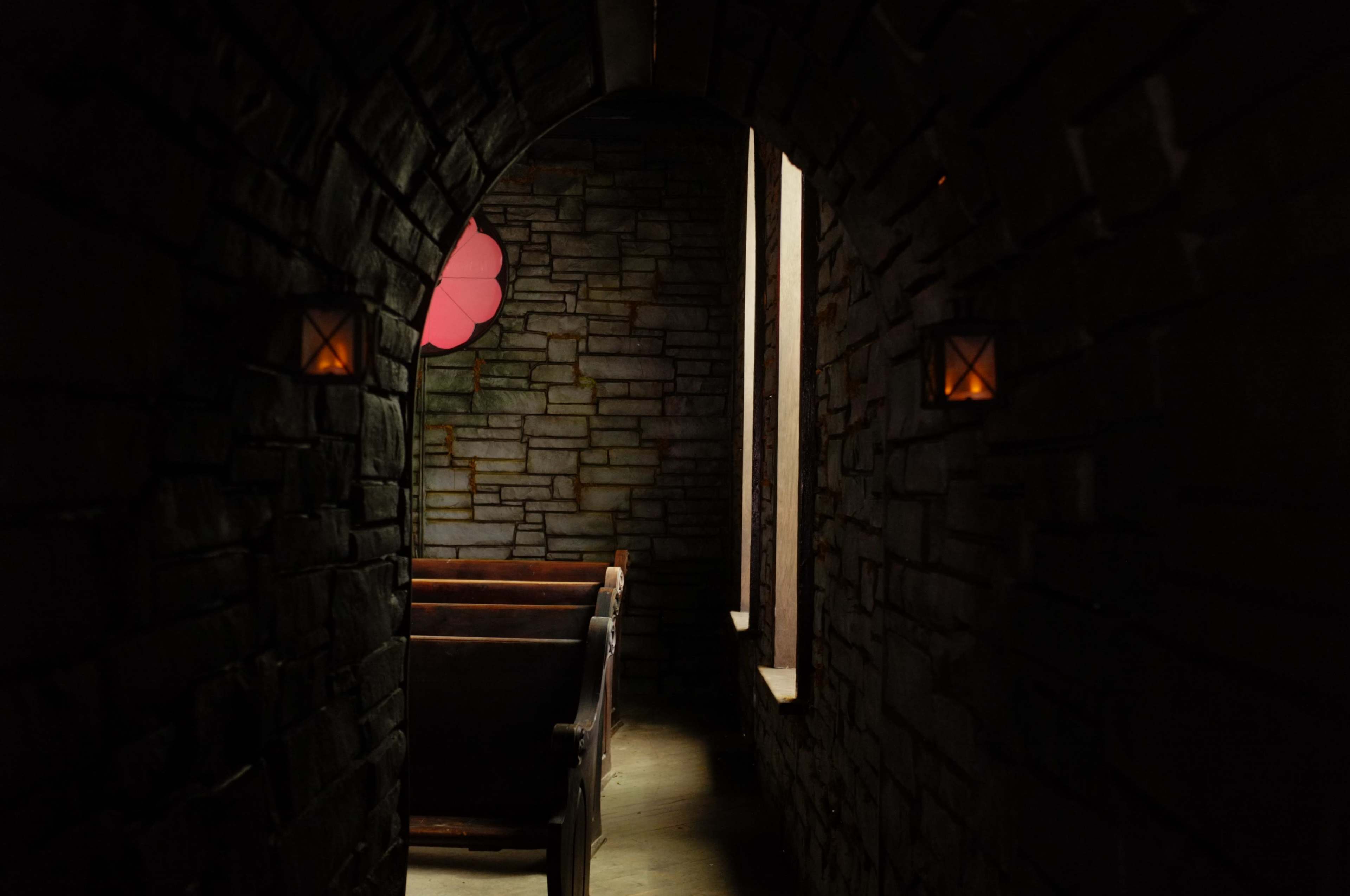 The image shows a dimly lit stone archway leading into a room with wooden pews and a colored window.