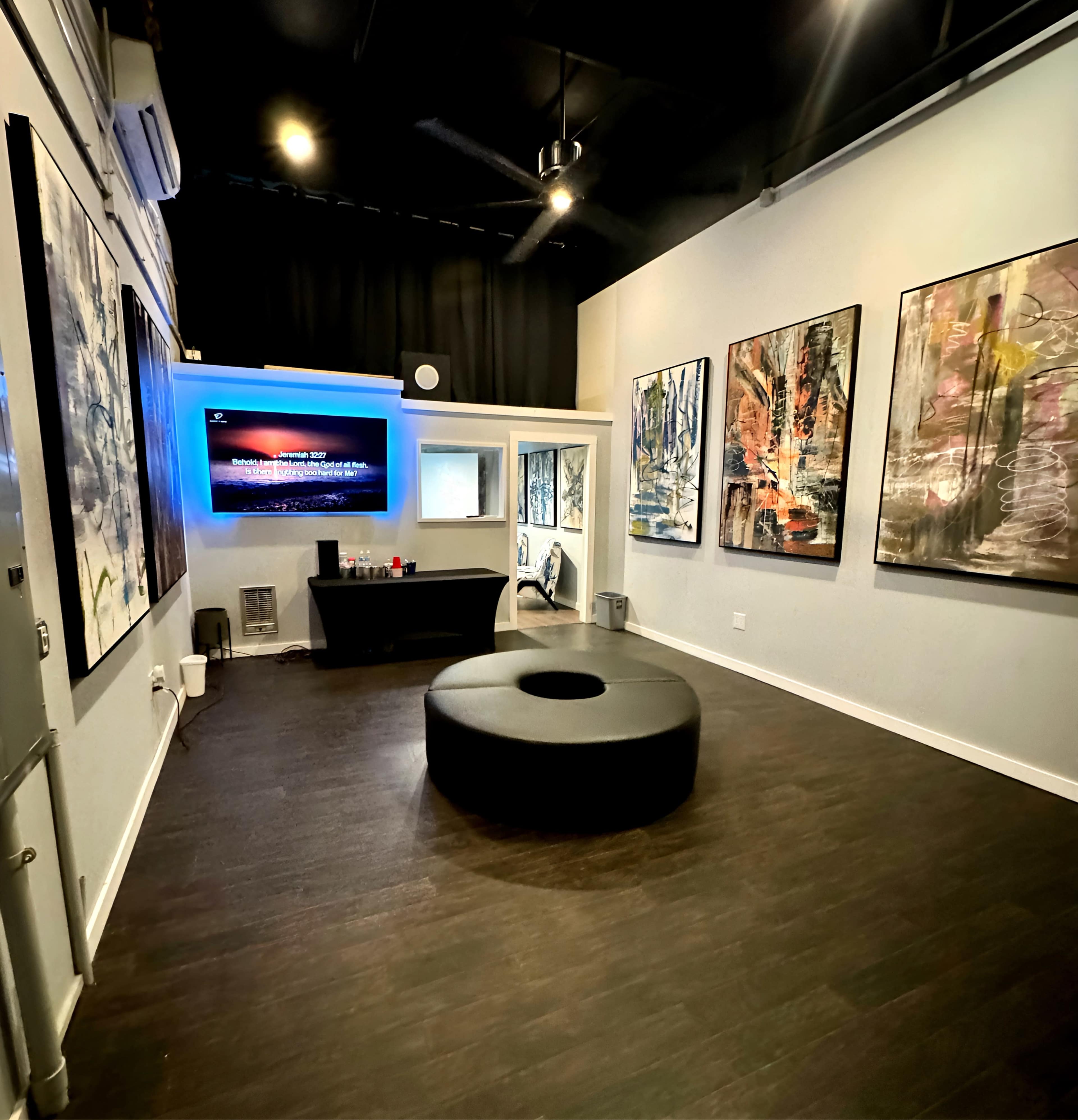 A modern art gallery with a circular seating area in the center, several abstract paintings on the walls, and a television mounted on the opposite wall.