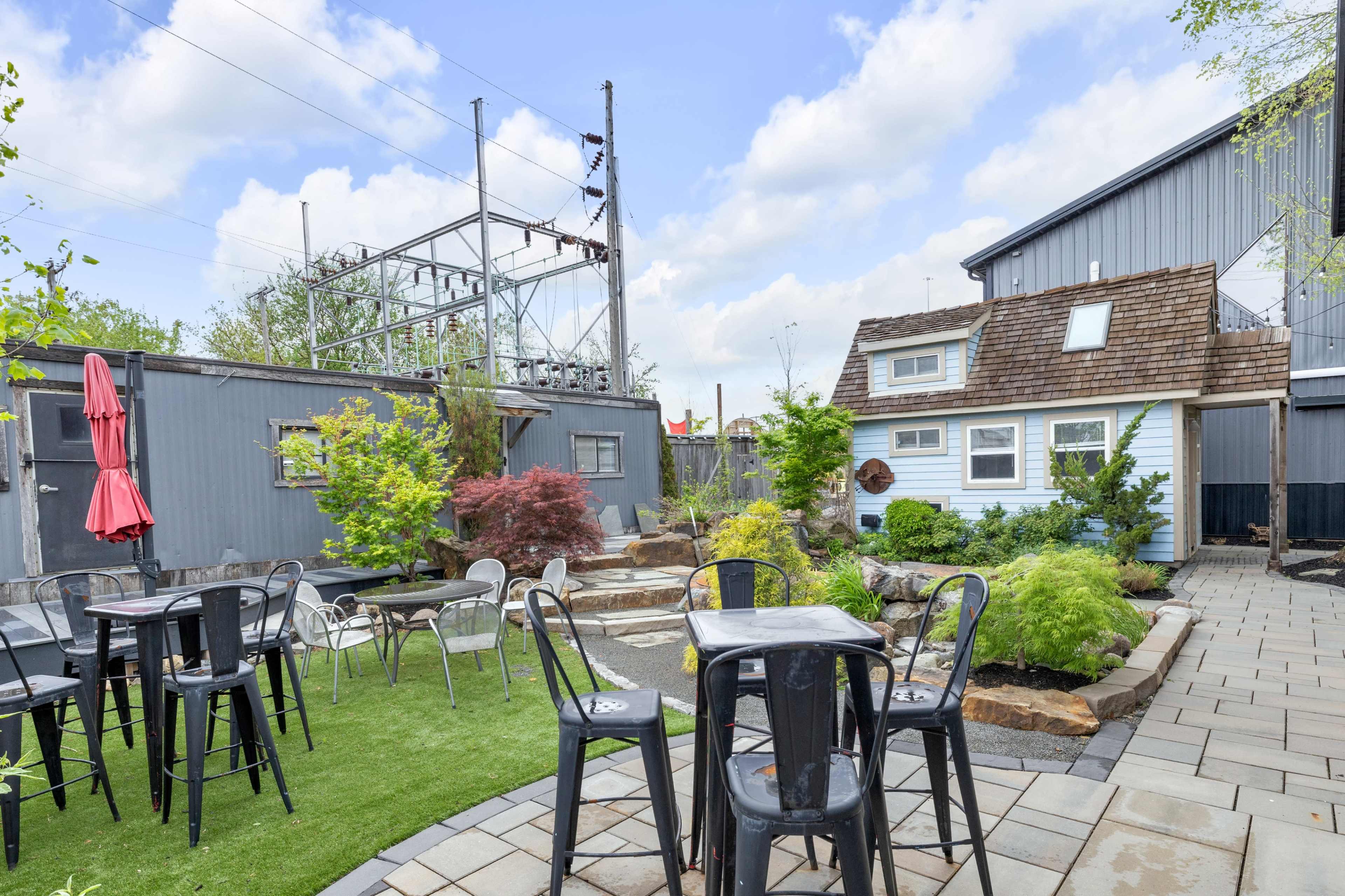 The image depicts a landscaped outdoor area featuring a small blue cottage, metal tables and chairs, and greenery, with electrical equipment visible in the background.