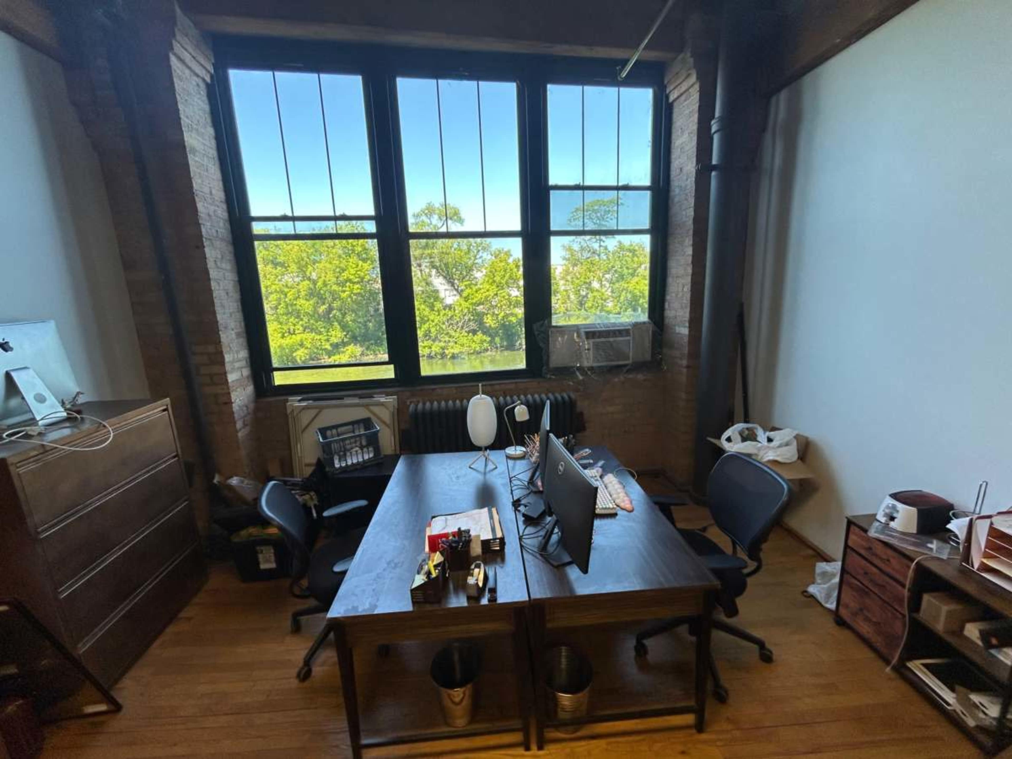 The image shows an office with two desks facing each other, large windows providing a view of trees outside, and minimal furnishings.