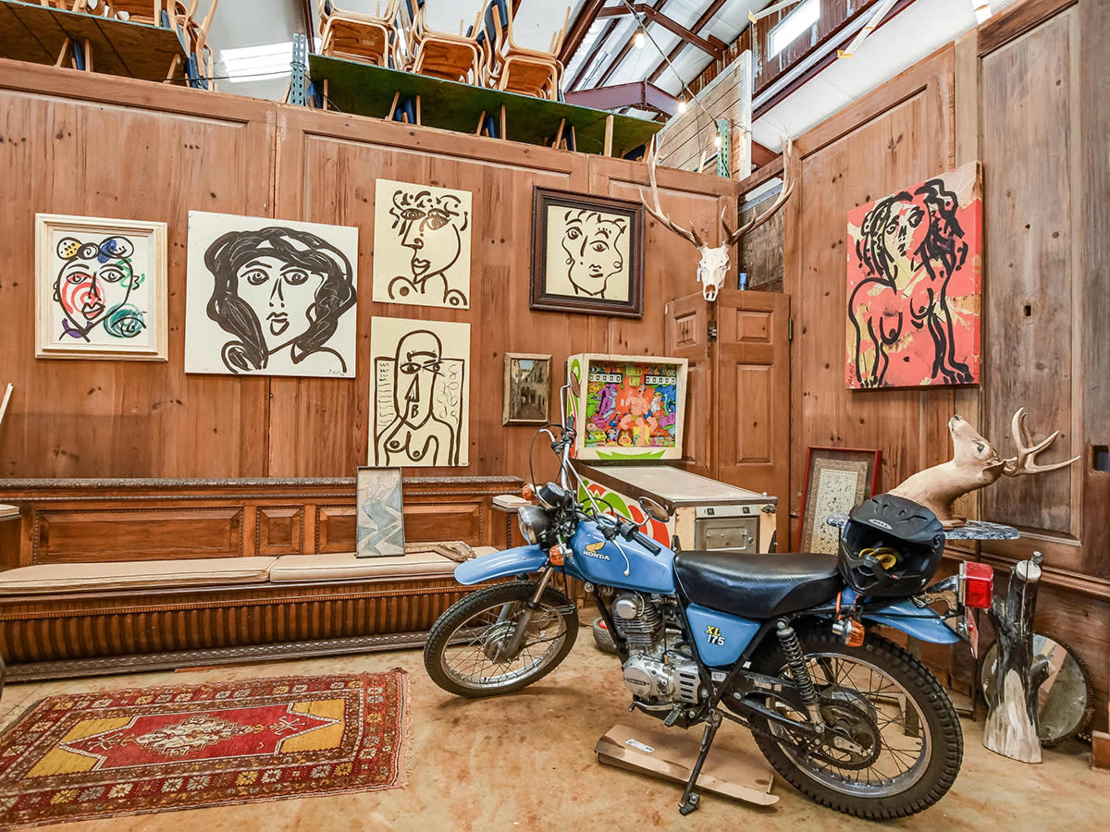 The scene features a blue motorcycle parked on a floor covered with a patterned rug, surrounded by wooden walls adorned with various artworks and mounted antlers.