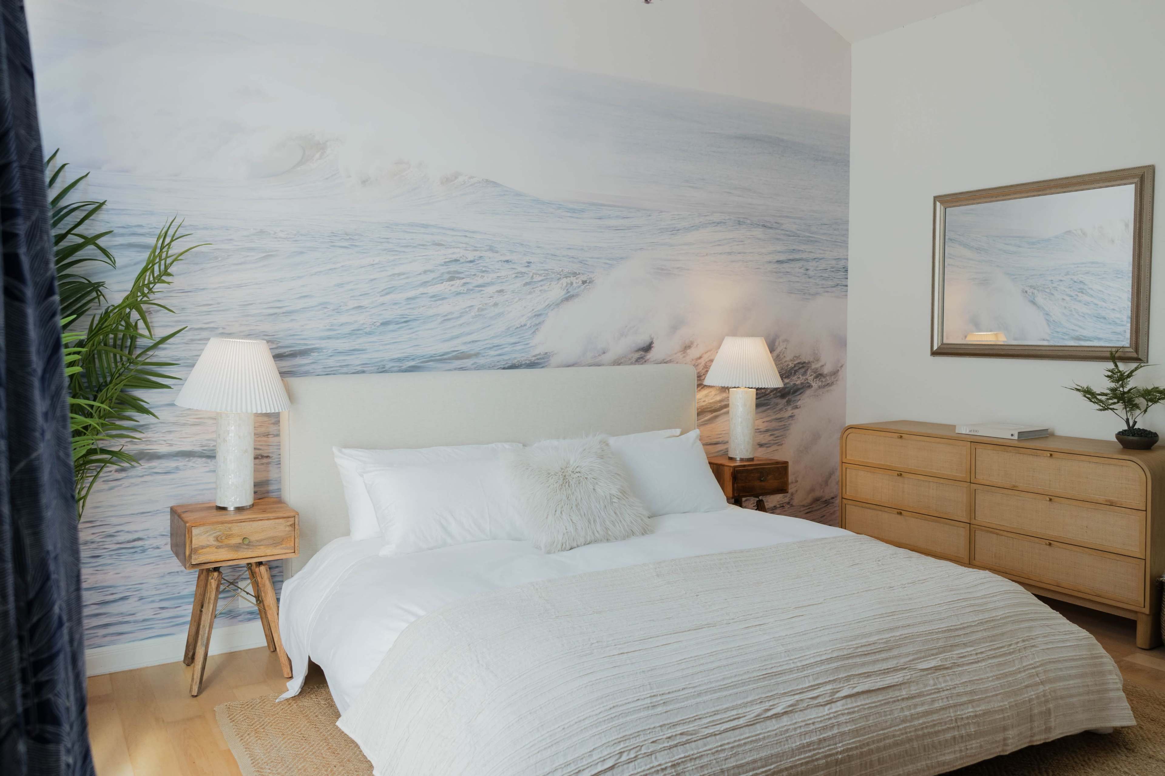The image shows a neatly arranged bedroom with a bed dressed in white linens, a wooden dresser, and a wall mural depicting ocean waves.