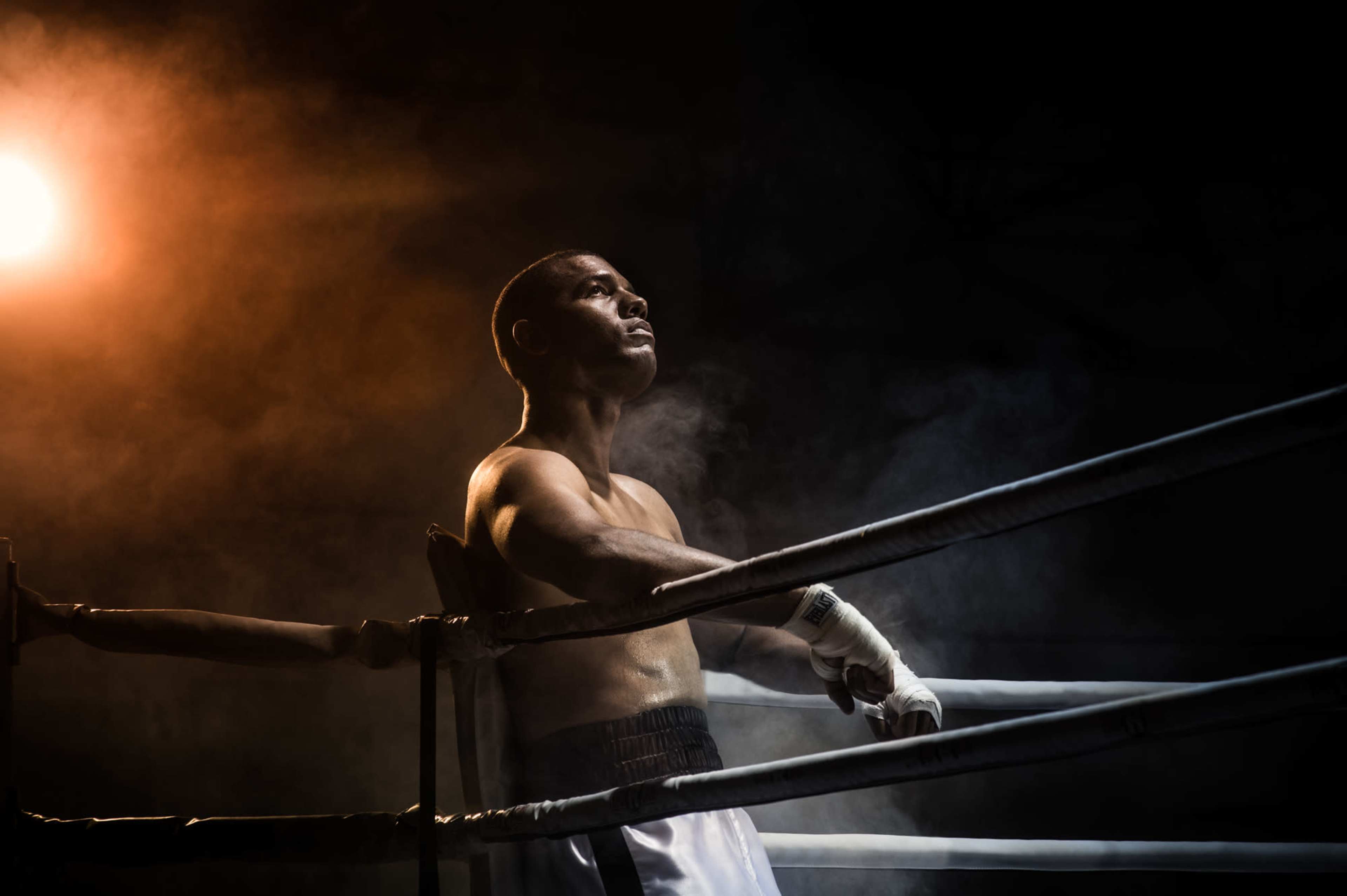 A boxer stands in a dimly lit ring, leaning against the ropes with a contemplative expression.