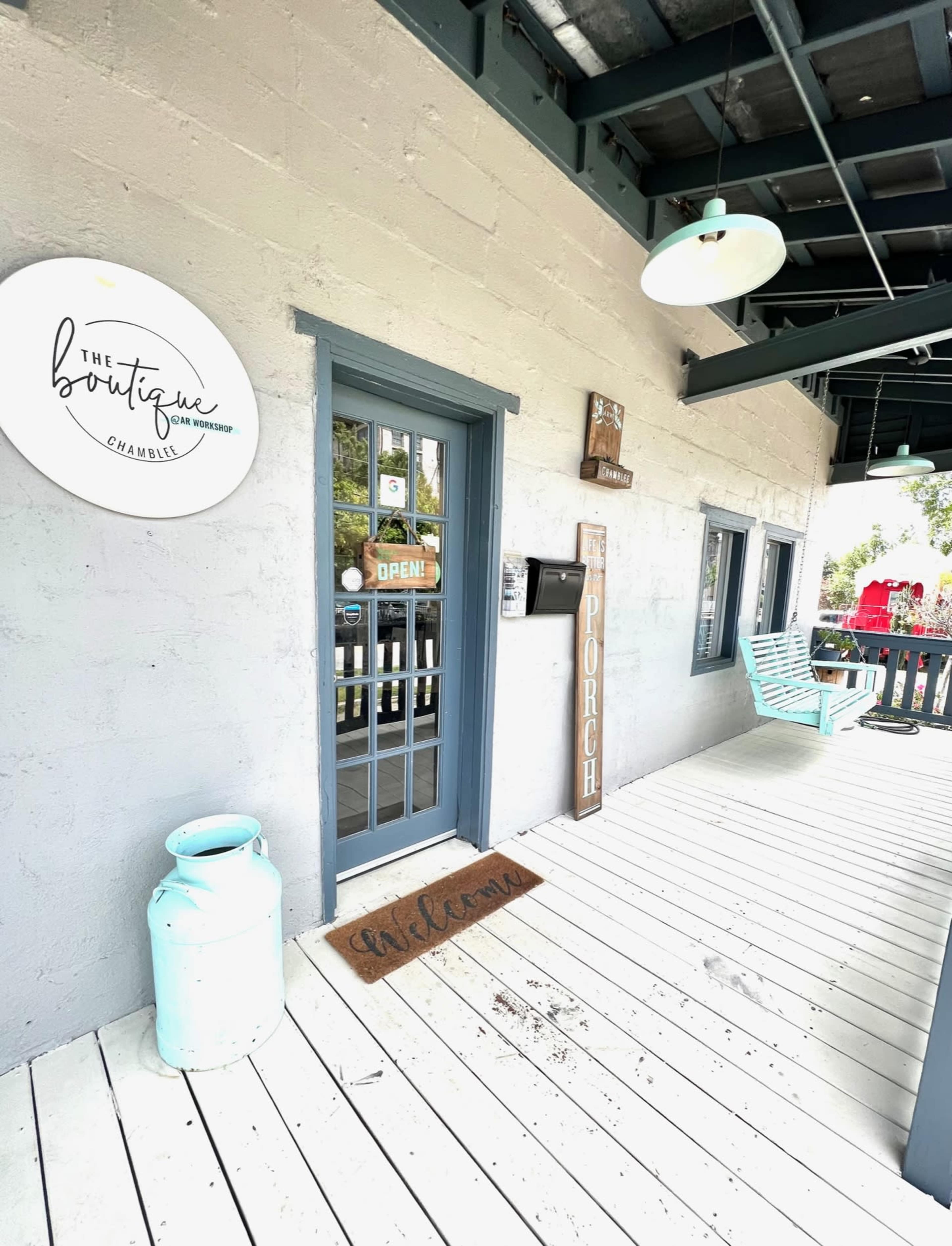 The entrance of a boutique called "The Boutique," featuring a door with a sign that reads "Open," a decorative porch with a blue bench, and a welcome mat.