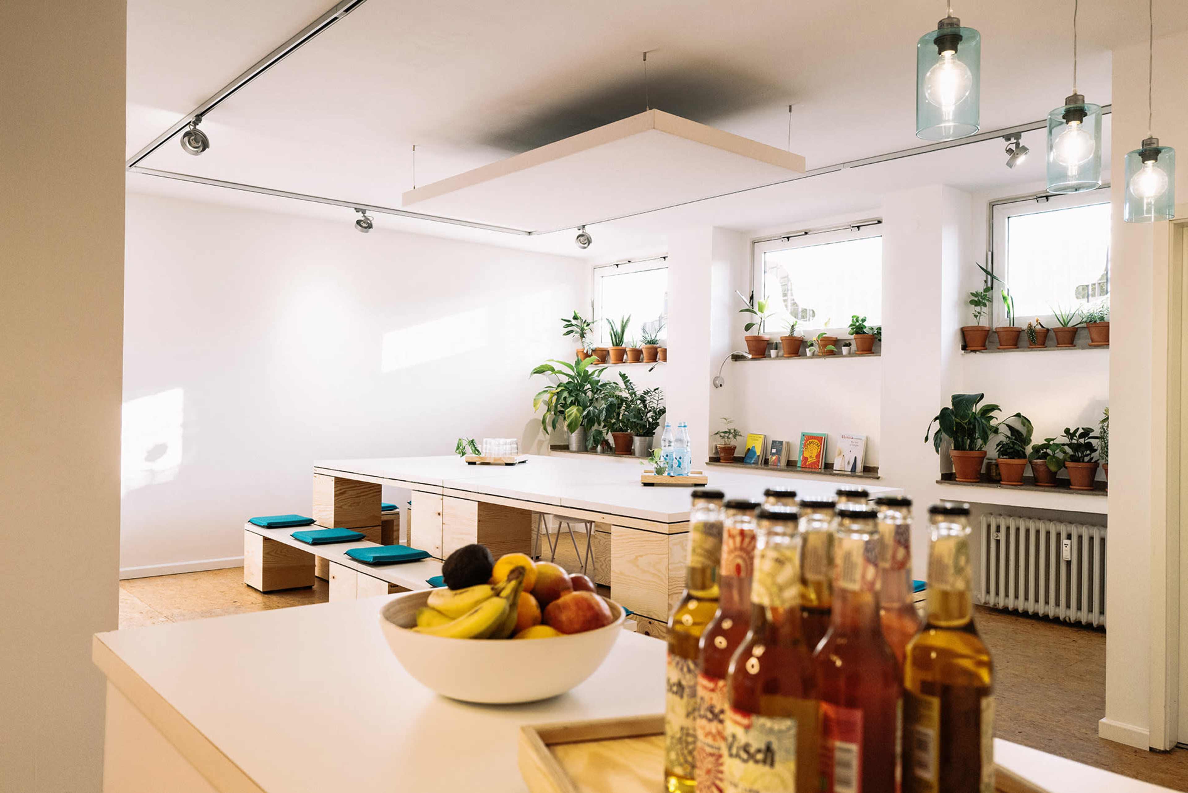 A bright room with wooden tables, green plants on shelves, and a bowl of fruit on a counter, along with bottles lined up on the edge.