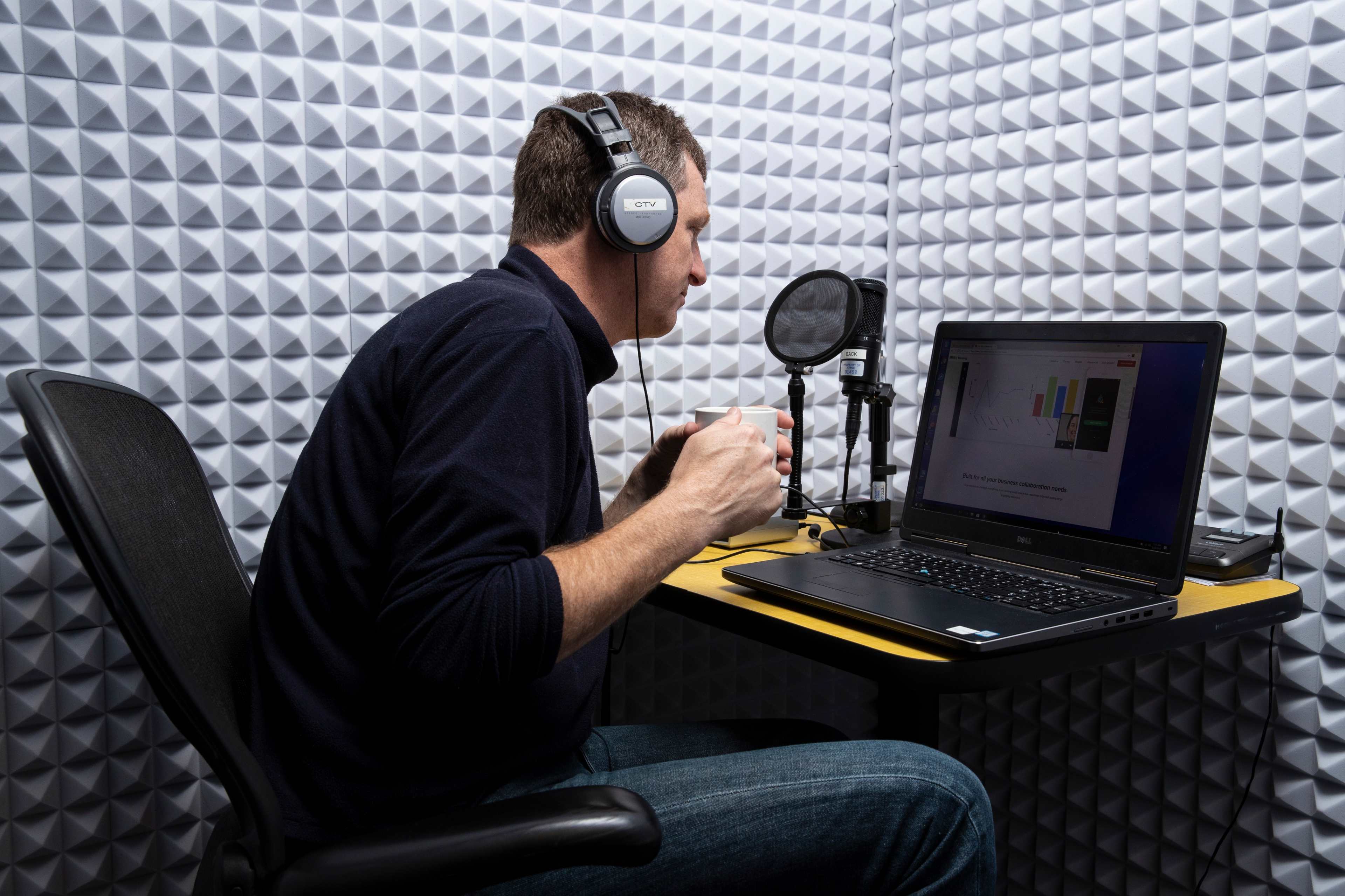 A person wearing headphones is sitting at a desk with a laptop and a microphone in a soundproof room.