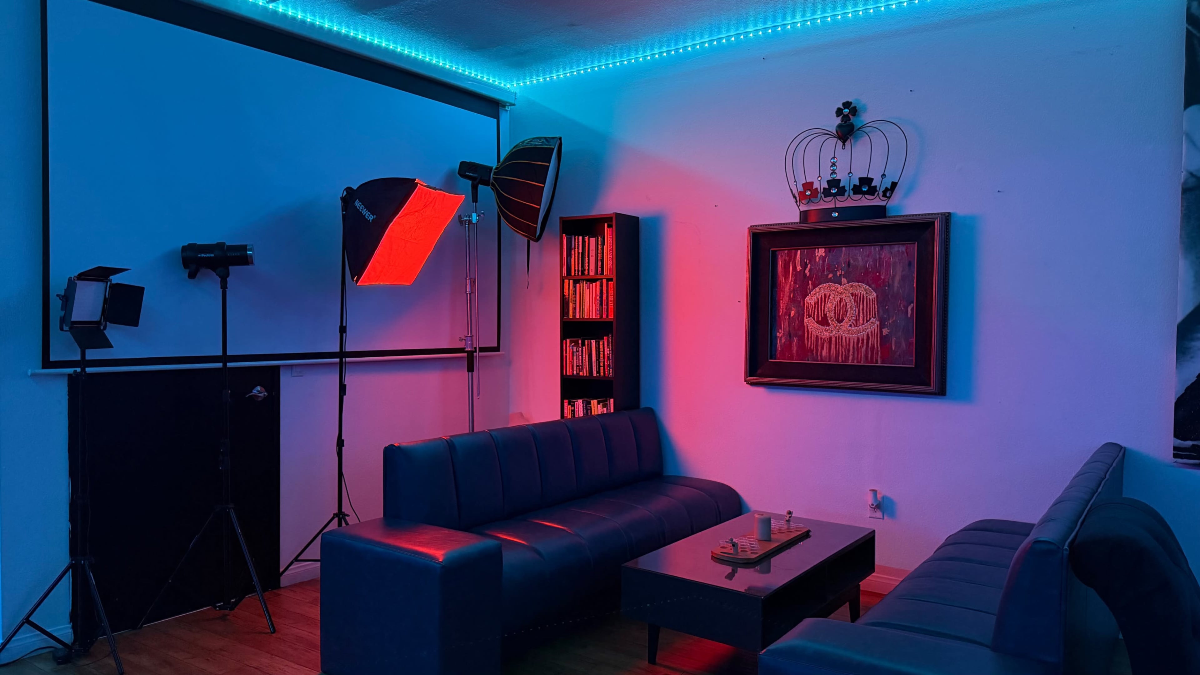The image shows a dimly lit room with blue and red lighting, featuring two dark sofas, a central coffee table, and a framed artwork above a shelf of books.