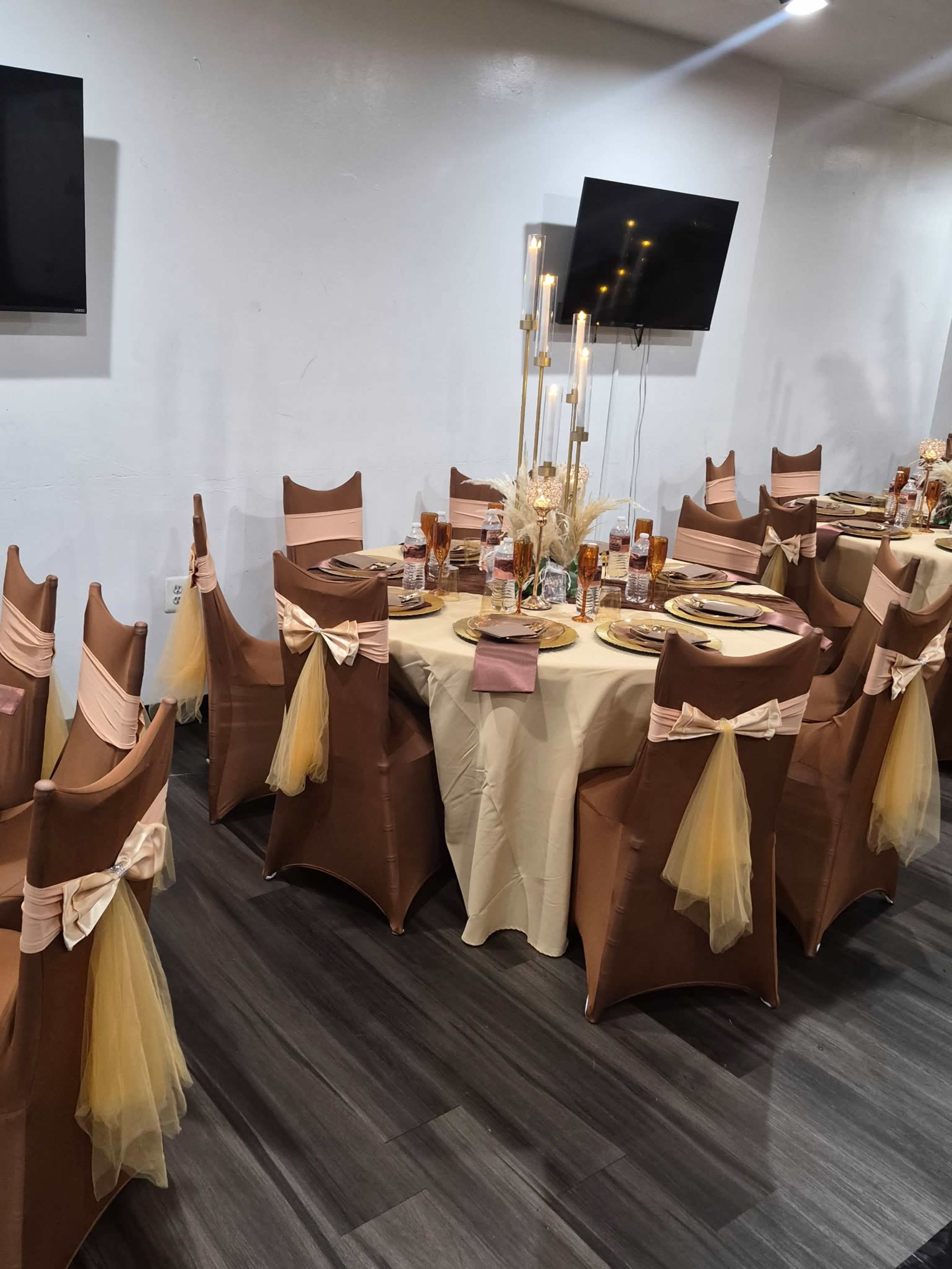 A round dining table is set with decorative plates and glassware, surrounded by chairs adorned with sashes, in a decorated event space.