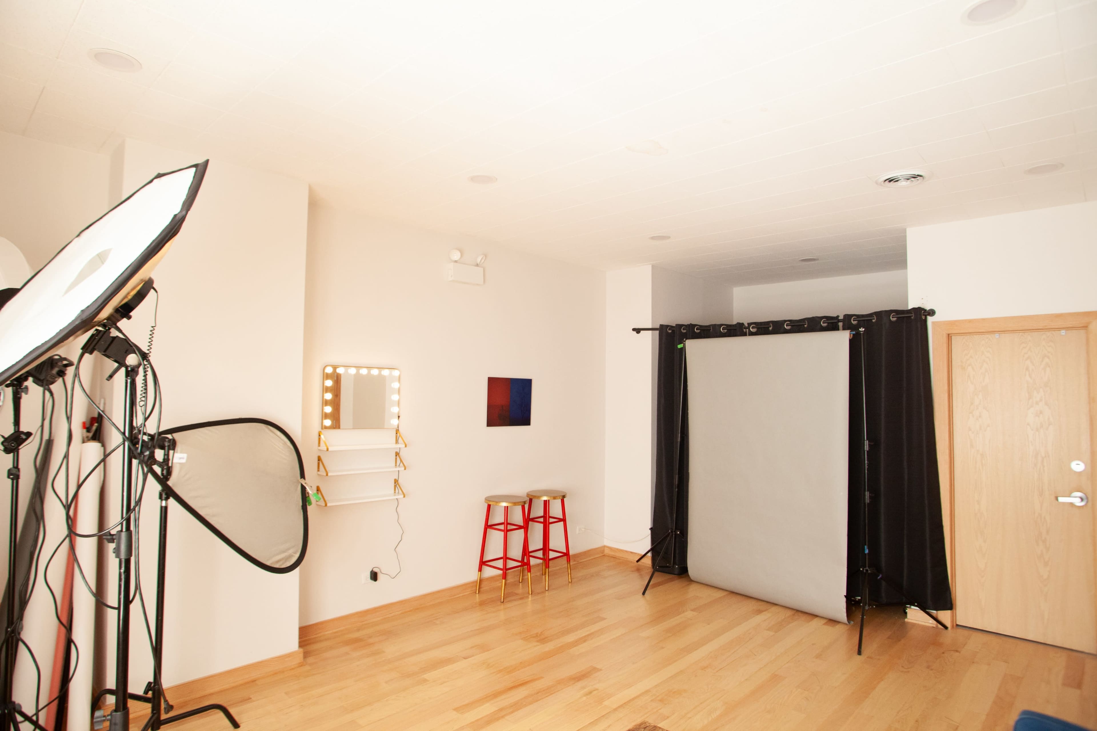 A bright photography studio with a gray backdrop, lighting equipment, and two stools positioned near the wall.