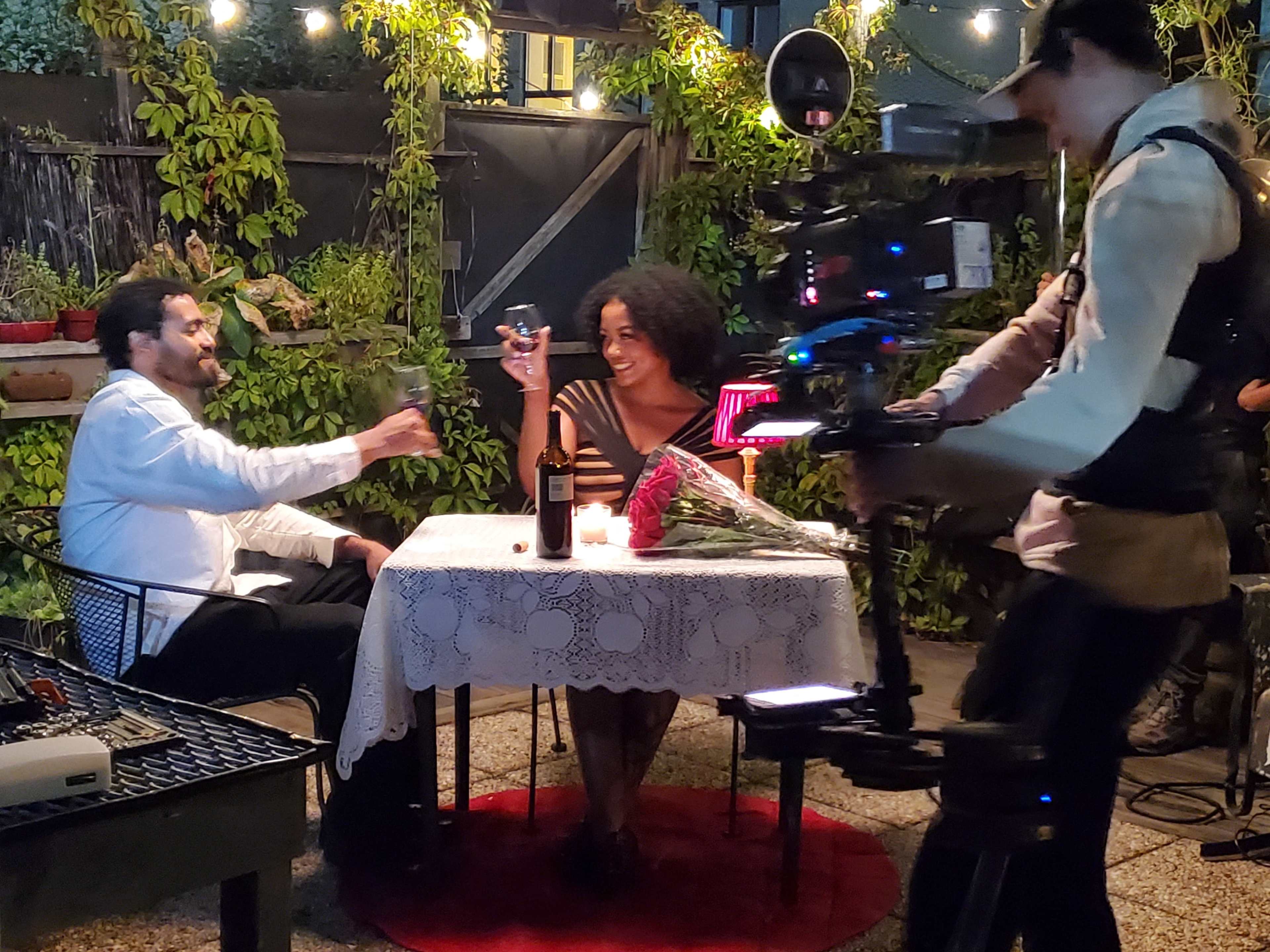 A couple sits at a table adorned with a tablecloth in a garden setting, raising glasses while a cameraman captures the moment.