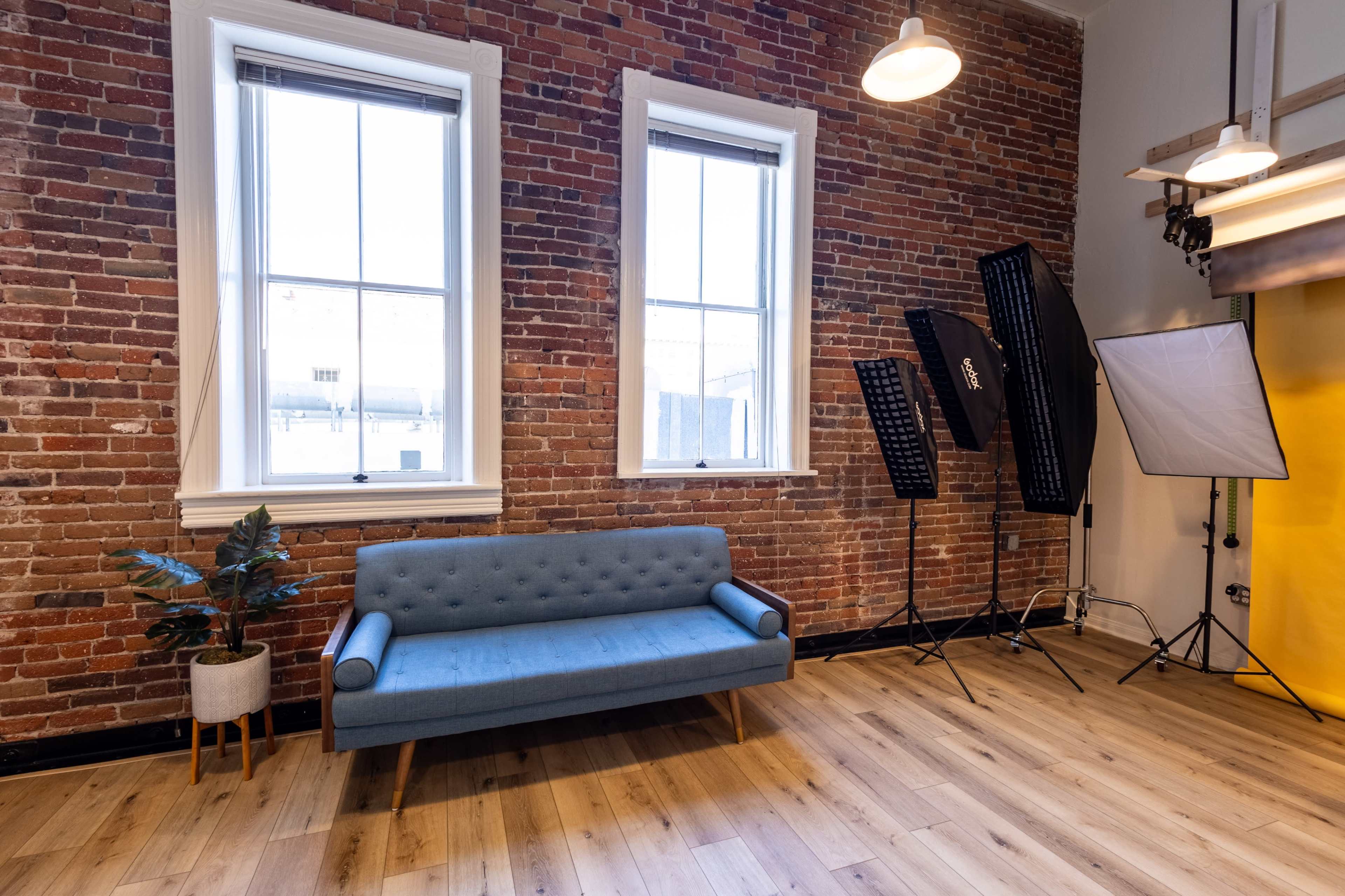 A blue tufted sofa sits against a brick wall with large windows, while photography equipment, including light stands and a backdrop, is arranged nearby.
