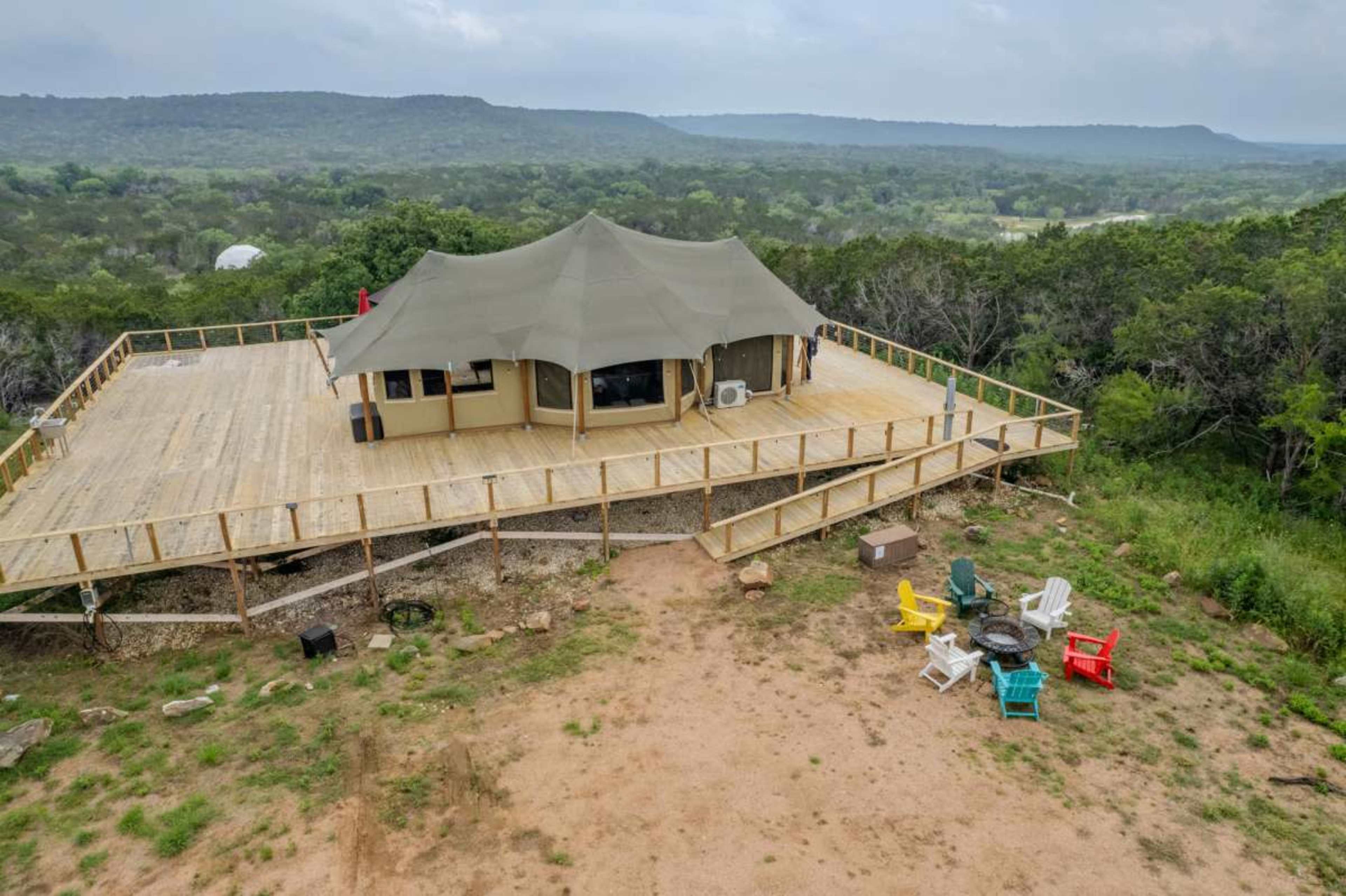 The image shows a large tent structure on a wooden deck surrounded by a natural landscape, with a circle of colorful chairs around a fire pit nearby.