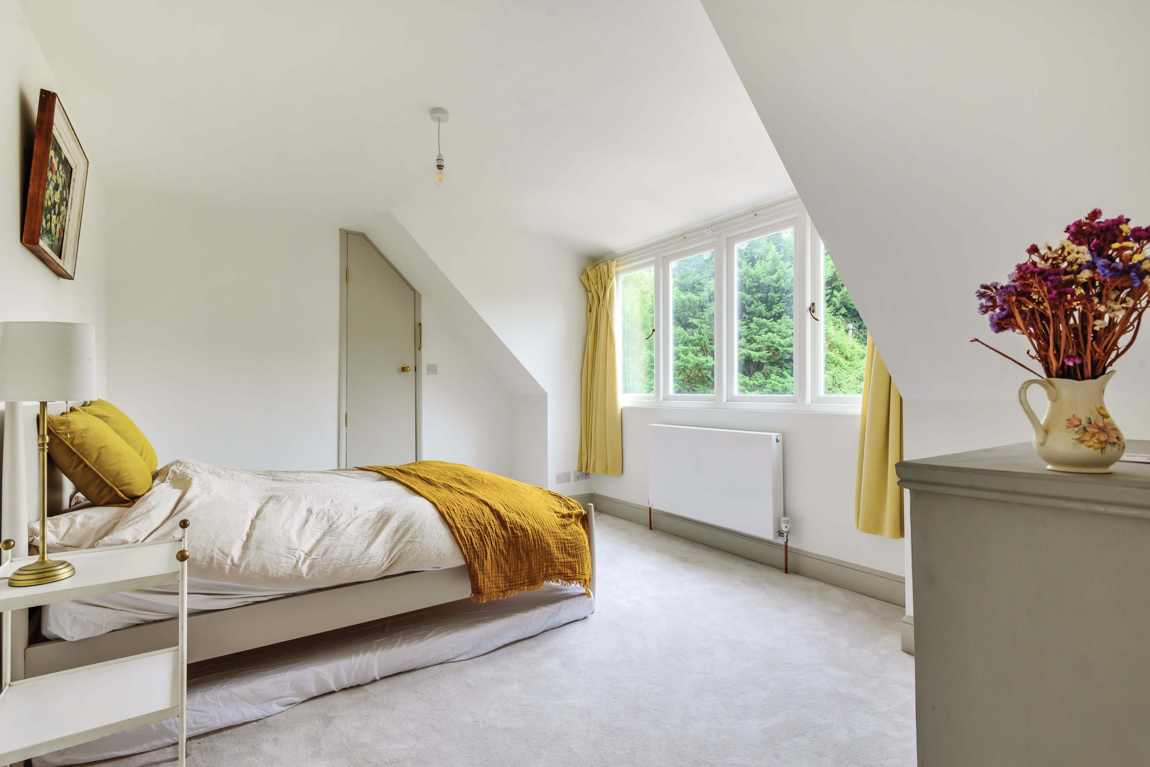 A brightly lit bedroom with a bed covered in a yellow blanket, large windows, and a vase of flowers on a dresser.