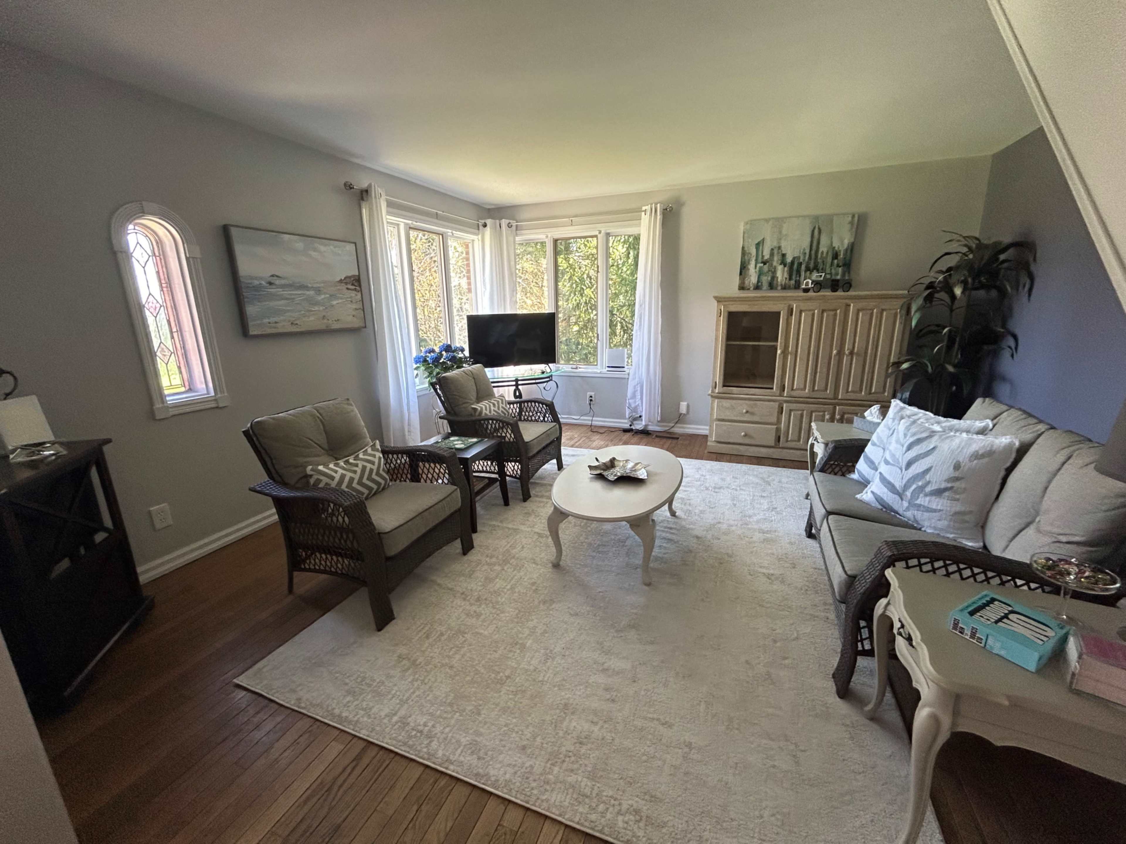 A living room features a light-colored rug, a small round table, two armchairs, and a sofa, all arranged around a window with natural light coming in.