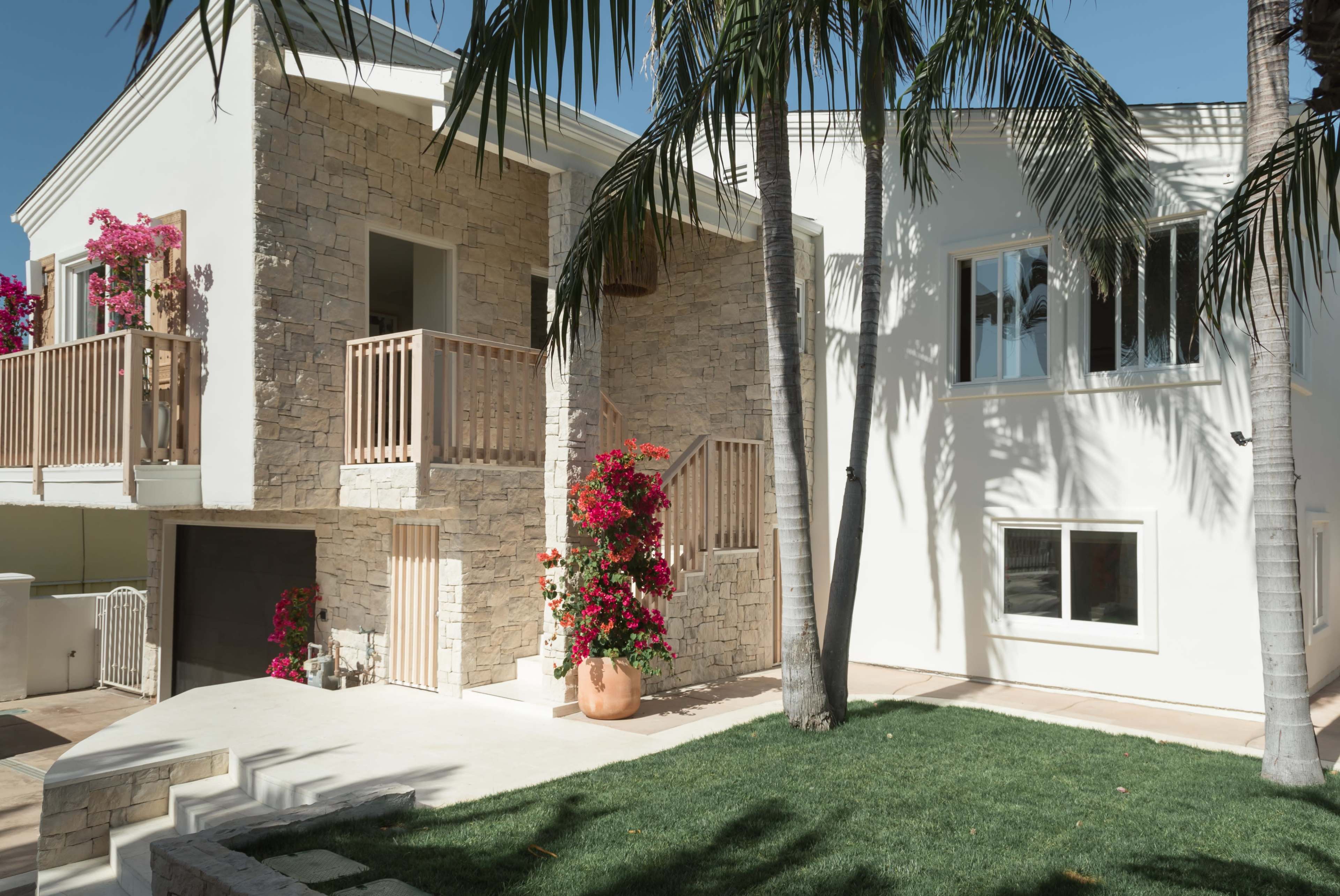 A modern two-story house with stone and white walls, featuring a balcony, palm trees, and vibrant flowers in large pots.