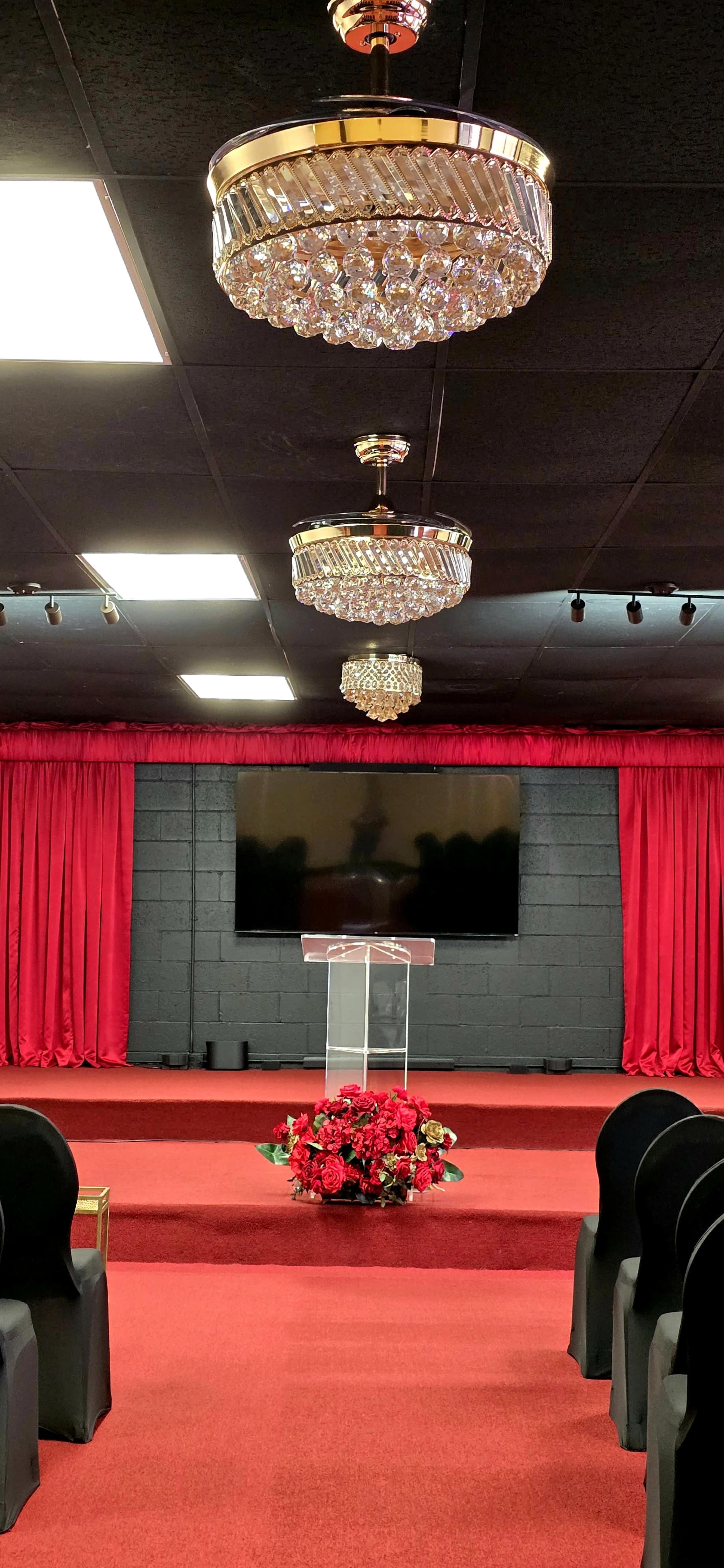 The image shows a decorated stage with a transparent podium, red curtains, black chairs, and chandeliers overhead in a ceremony space.
