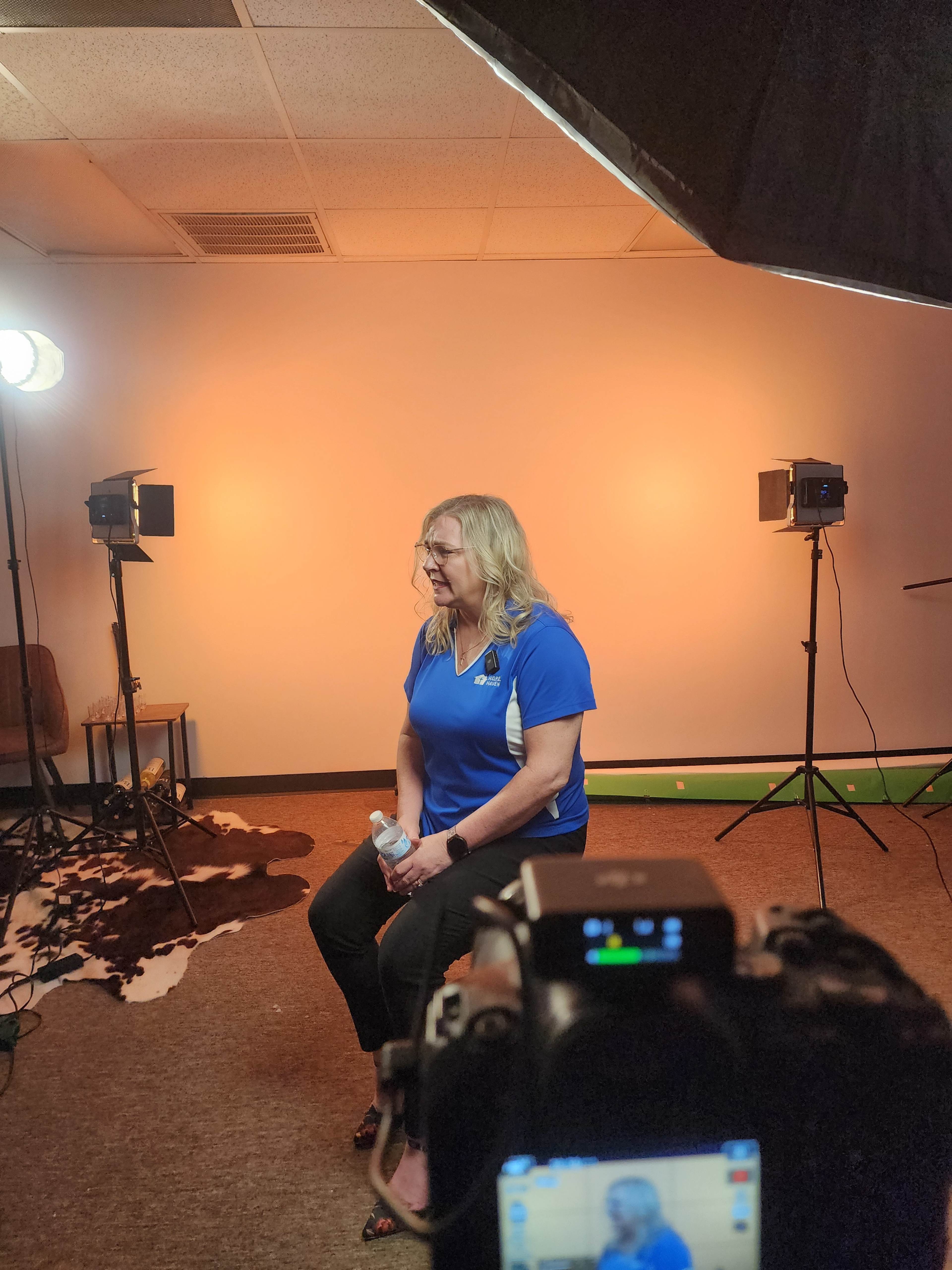 A woman in a blue shirt sits on a chair in a studio with soft lighting and cameras set up around her.