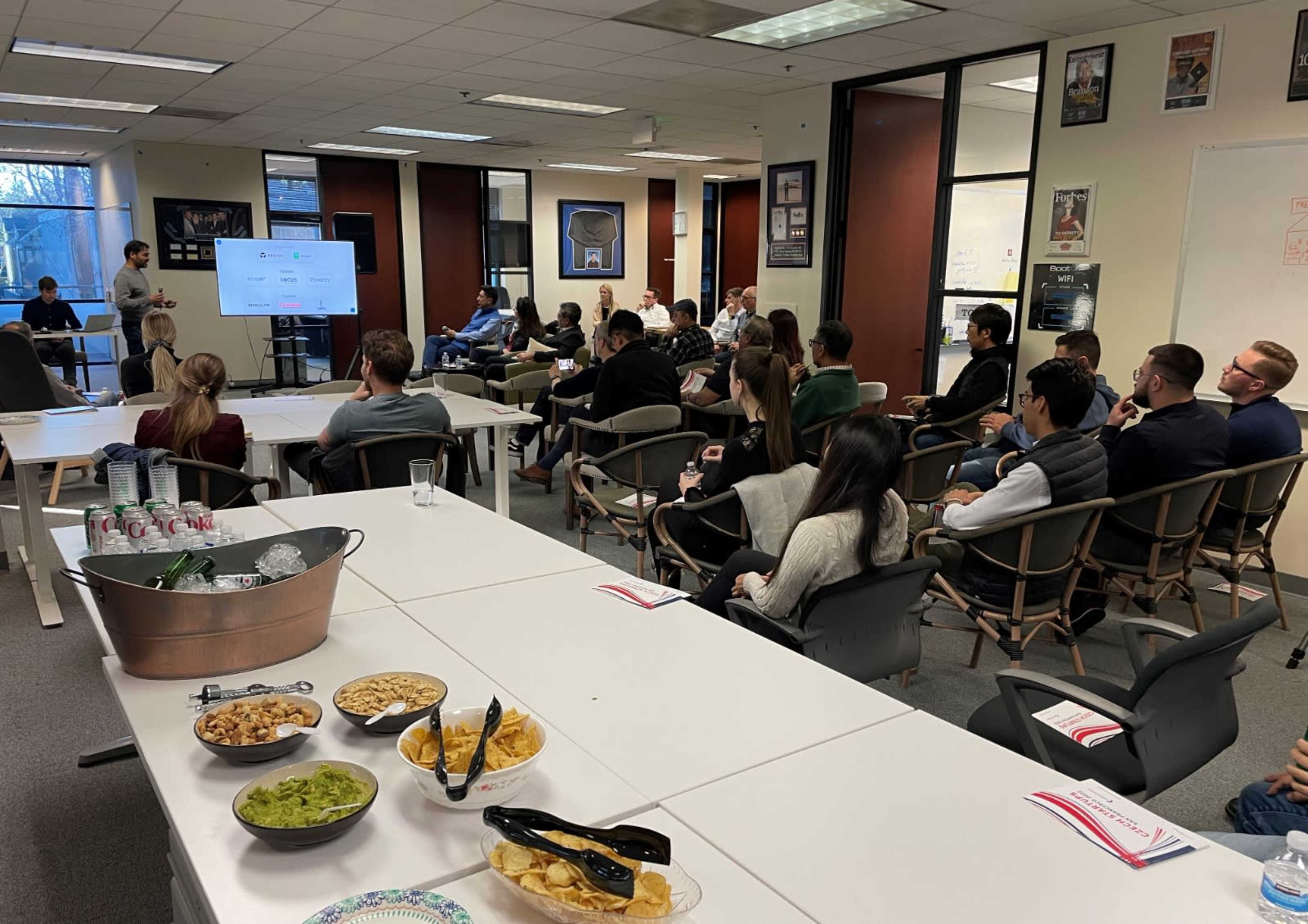 A group of people is seated in a meeting room, attentively watching a presentation while snacks and beverages are arranged on a table.
