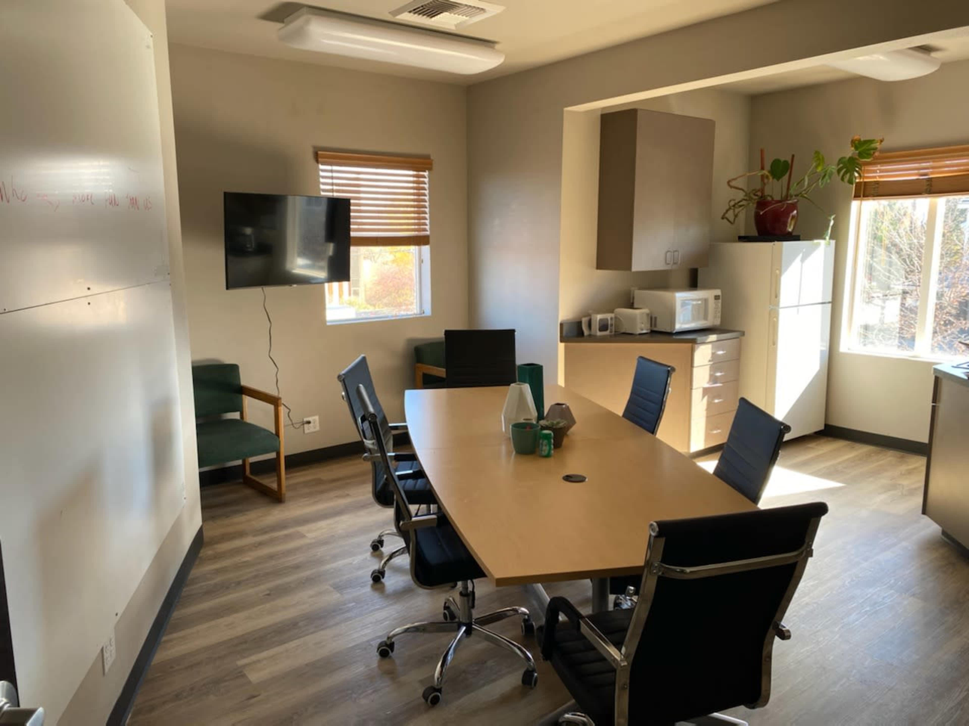 A conference room with a large table surrounded by black chairs, a television mounted on the wall, and a kitchenette in the background.
