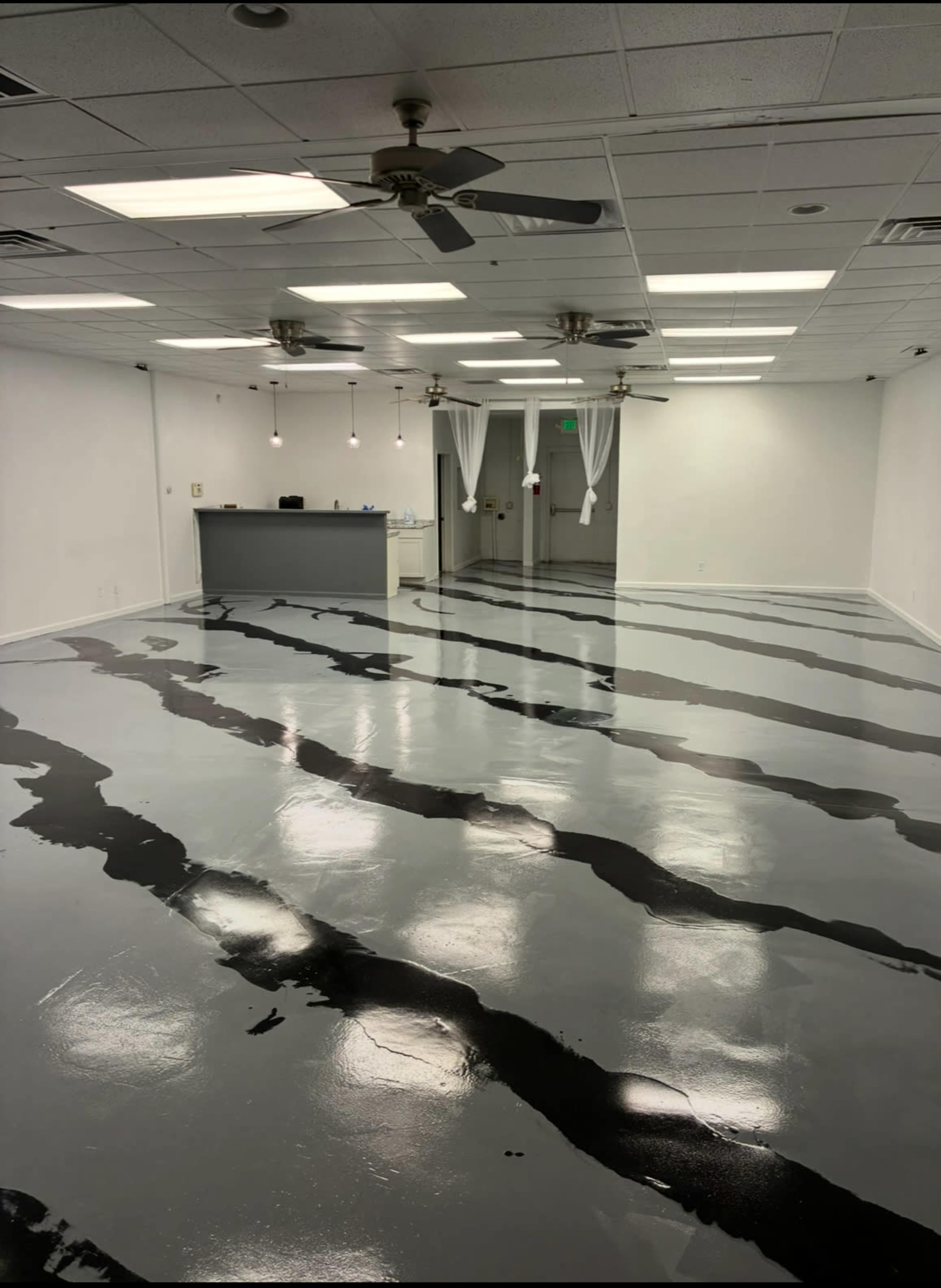 The image shows an empty room with a gray and black striped floor, a reception area in the back, and several ceiling fans.