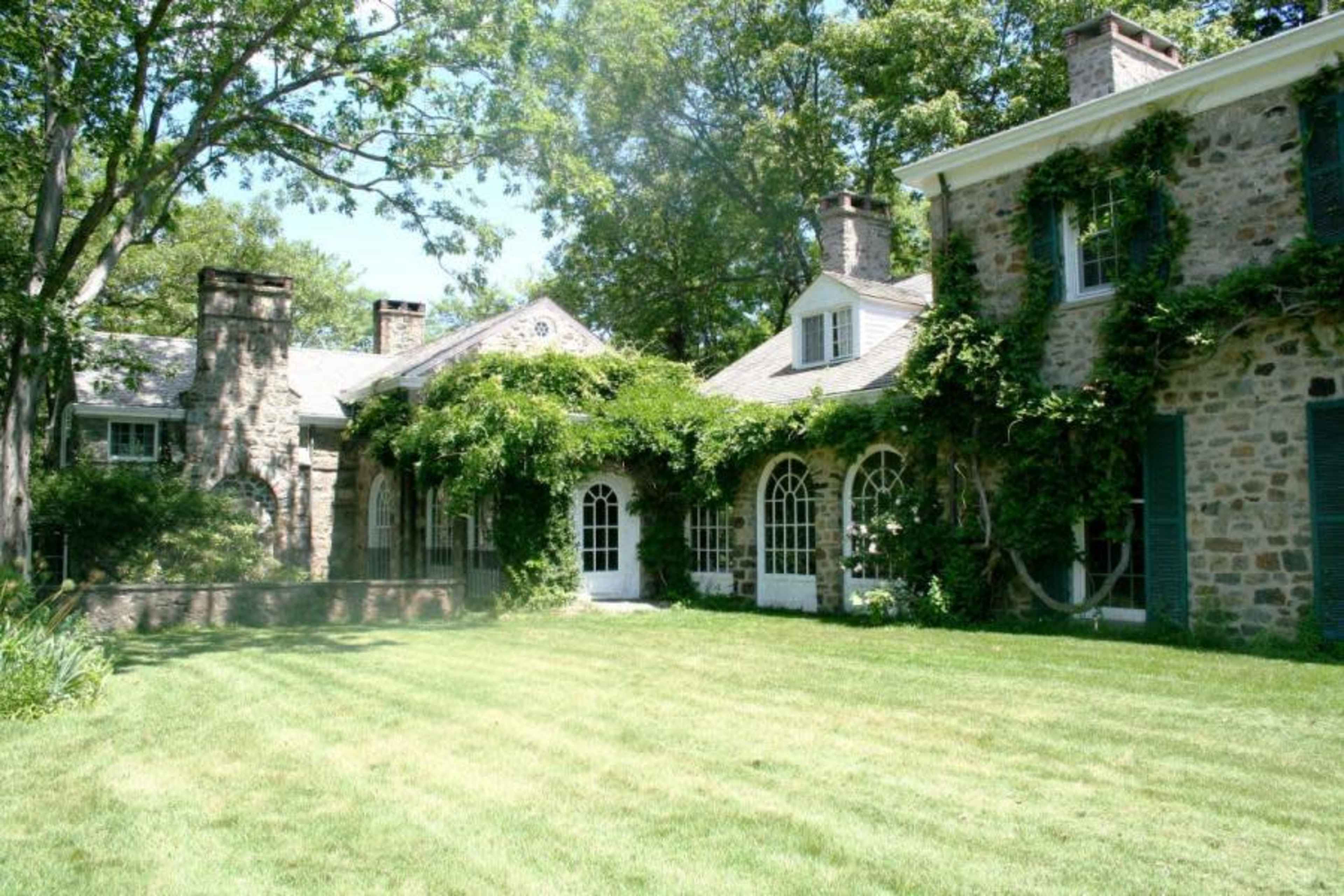 The image shows a stone house with large windows, surrounded by greenery and a well-manicured lawn.