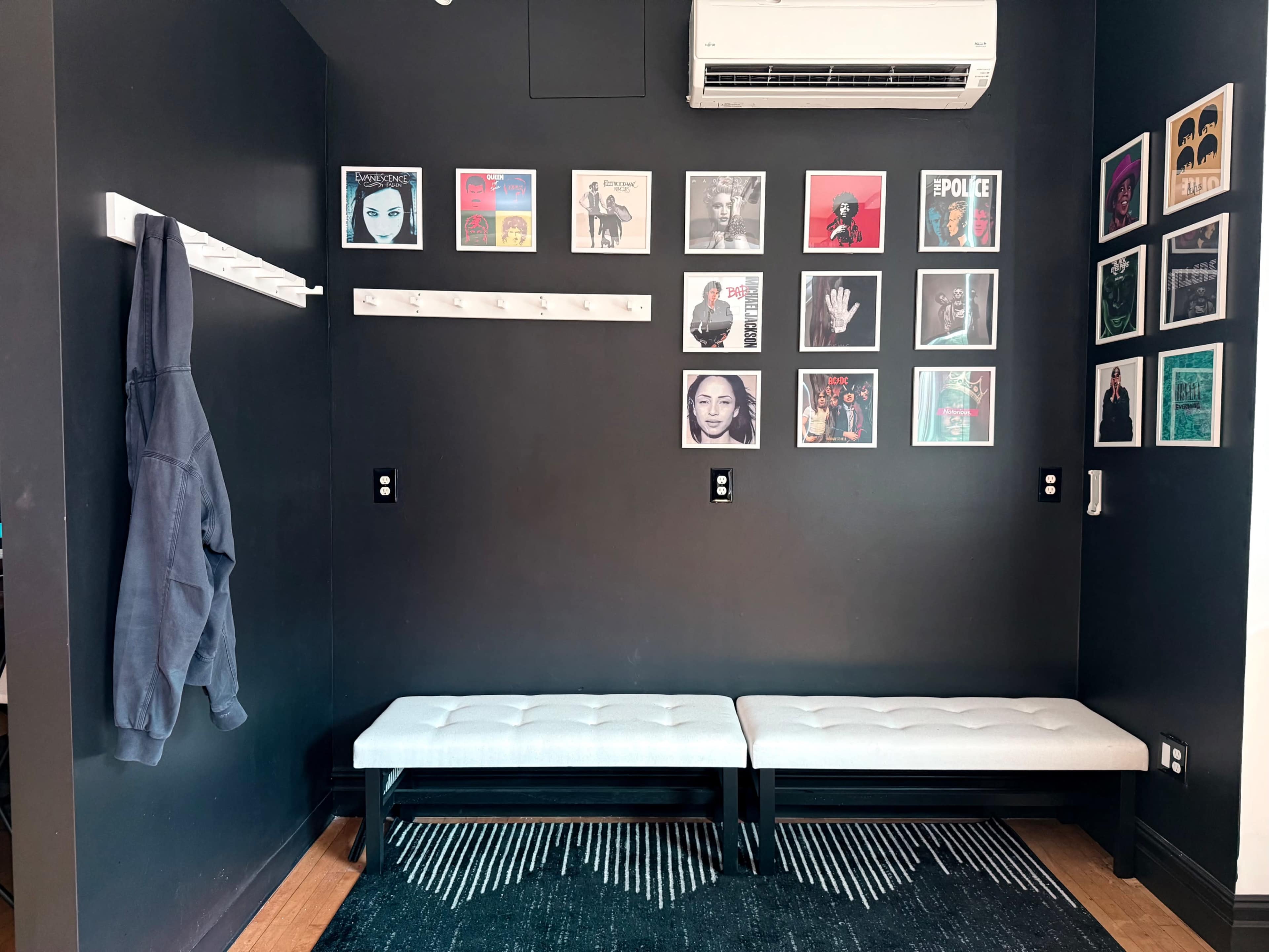 The image shows a sleek, modern entryway with black walls, two benches, a hook for hanging a hoodie, and framed artwork displayed on the walls.