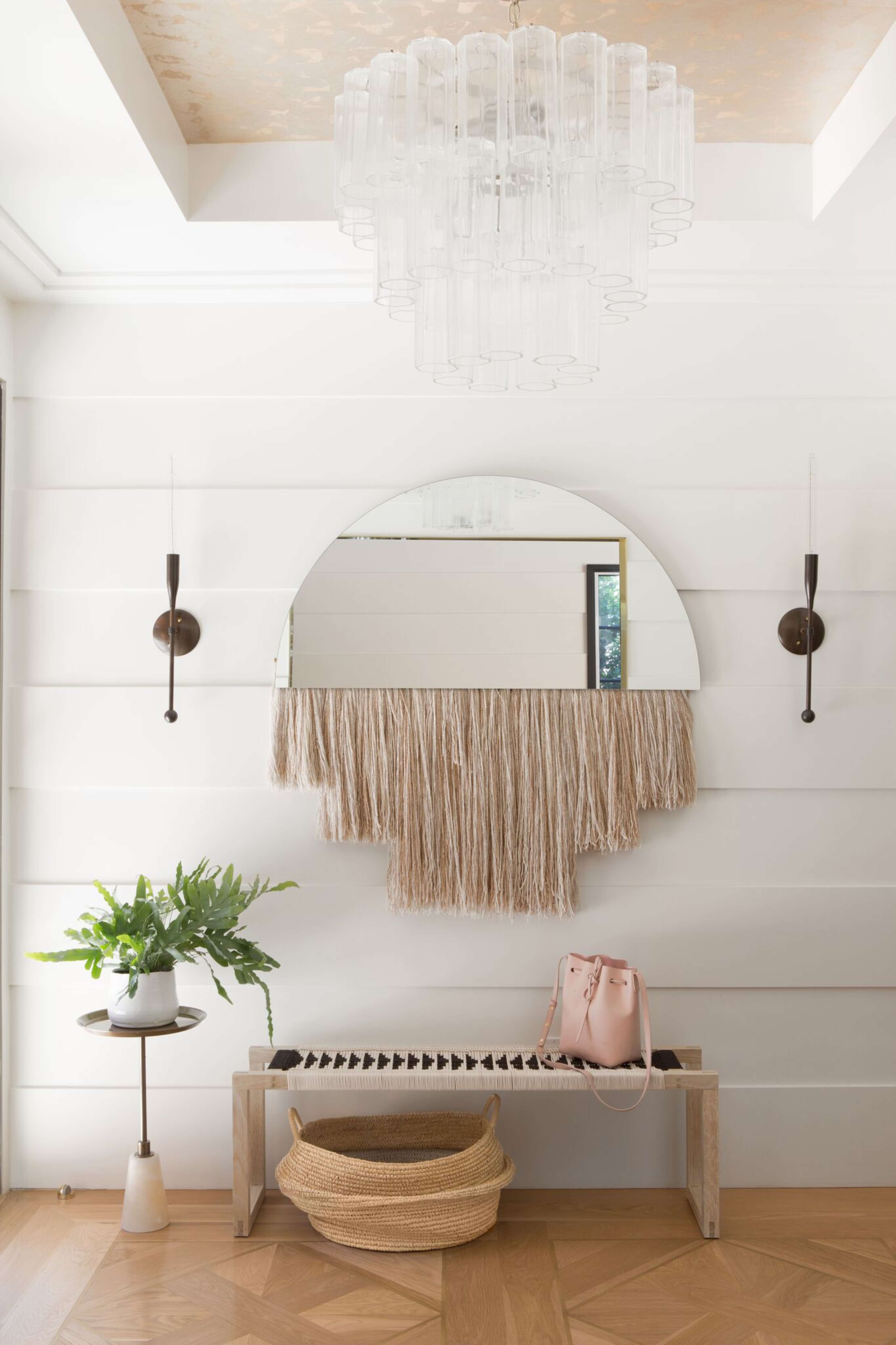 A modern entryway with a circular mirror featuring a fringe design, a stylish chandelier, a wall-mounted sconce, a small table with a plant, and a bench with a handbag on it.
