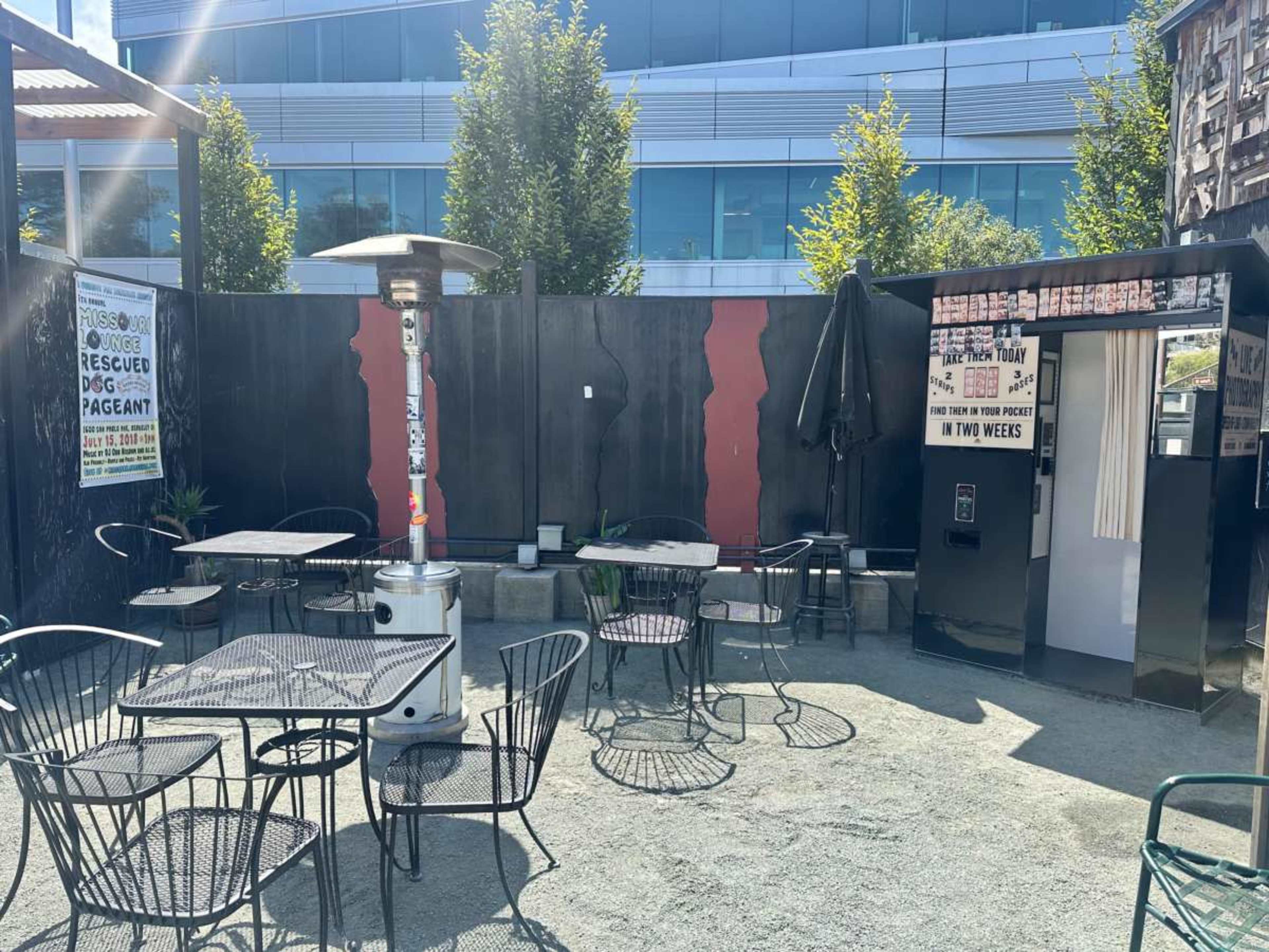 The image shows an outdoor seating area with black metal tables and chairs, surrounded by a black wall and a few trees, featuring a heater and a small booth.