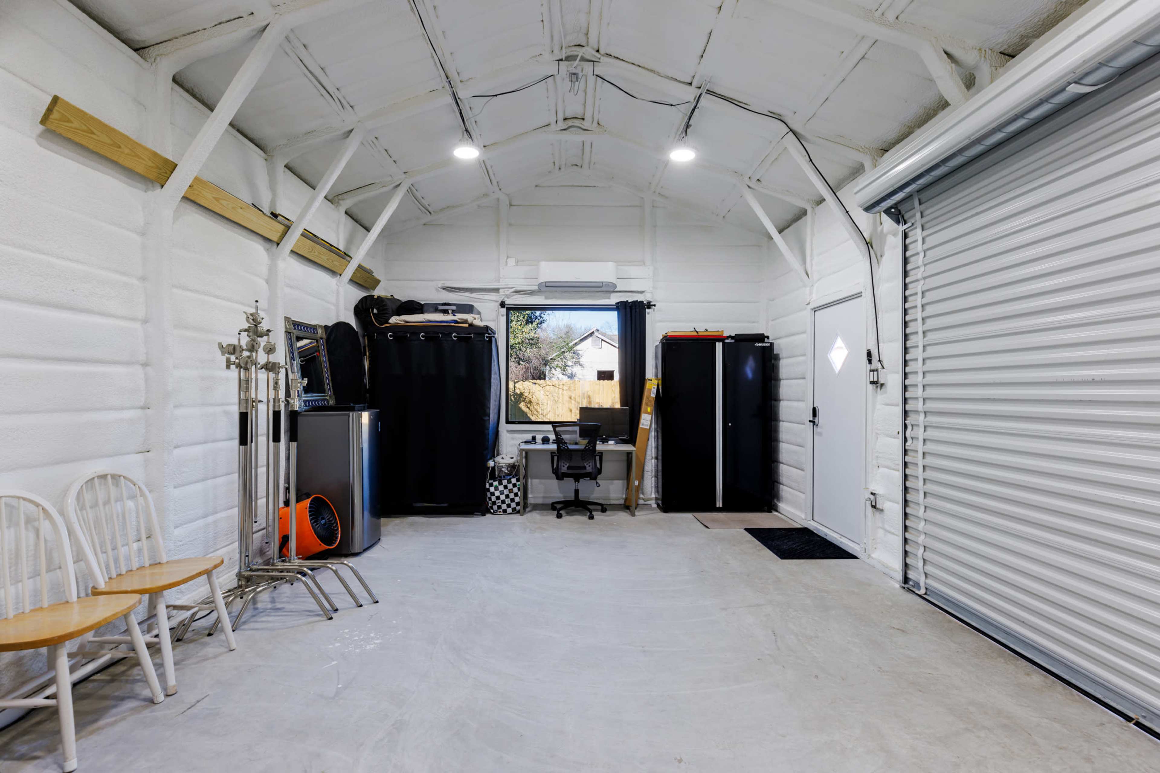 A white interior of a spacious garage with a desk and chair, a black refrigerator, and a few wooden chairs arranged against one wall.