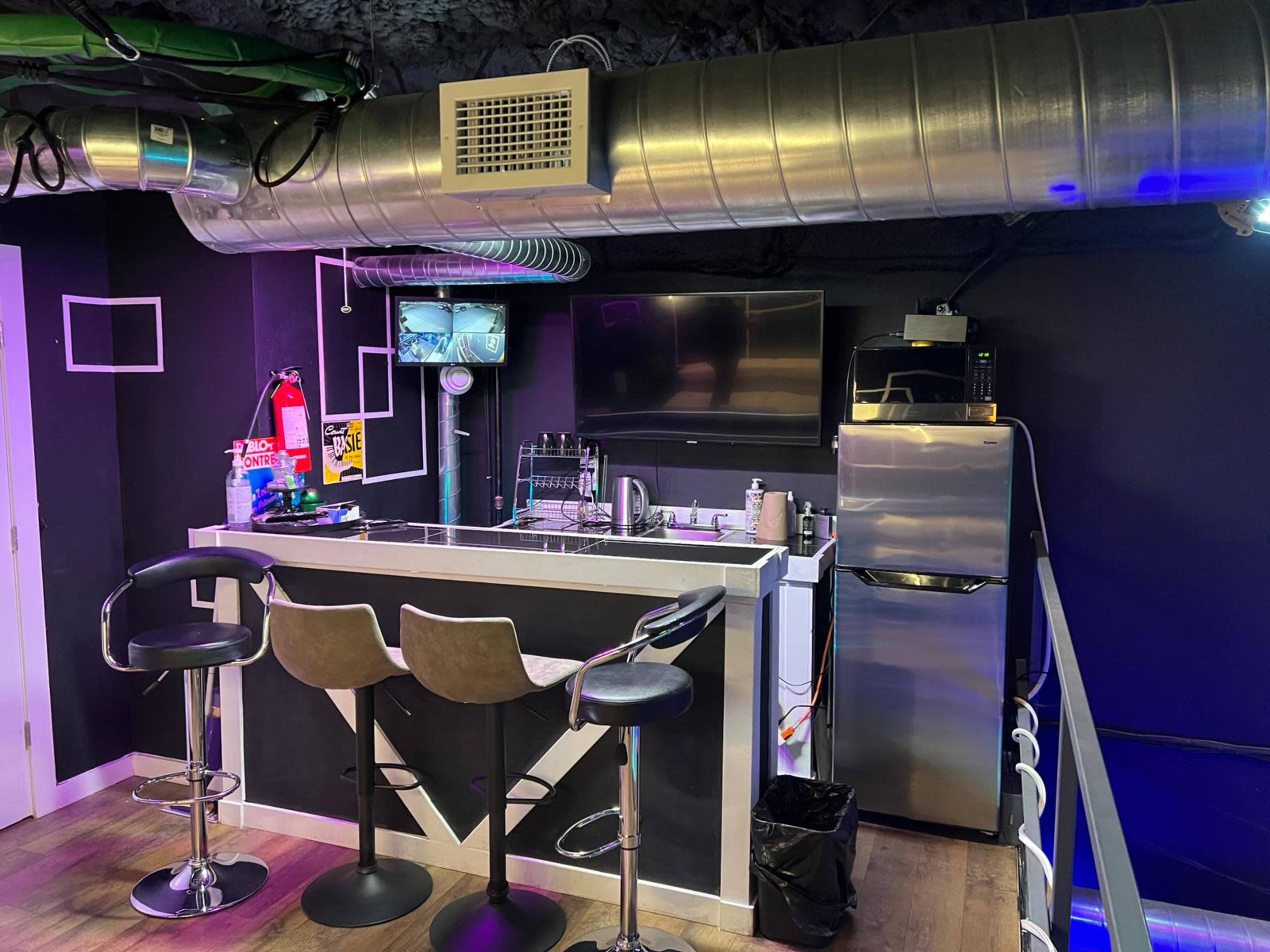 A modern bar area with two stools, a countertop, a television, and various appliances against a dark wall.