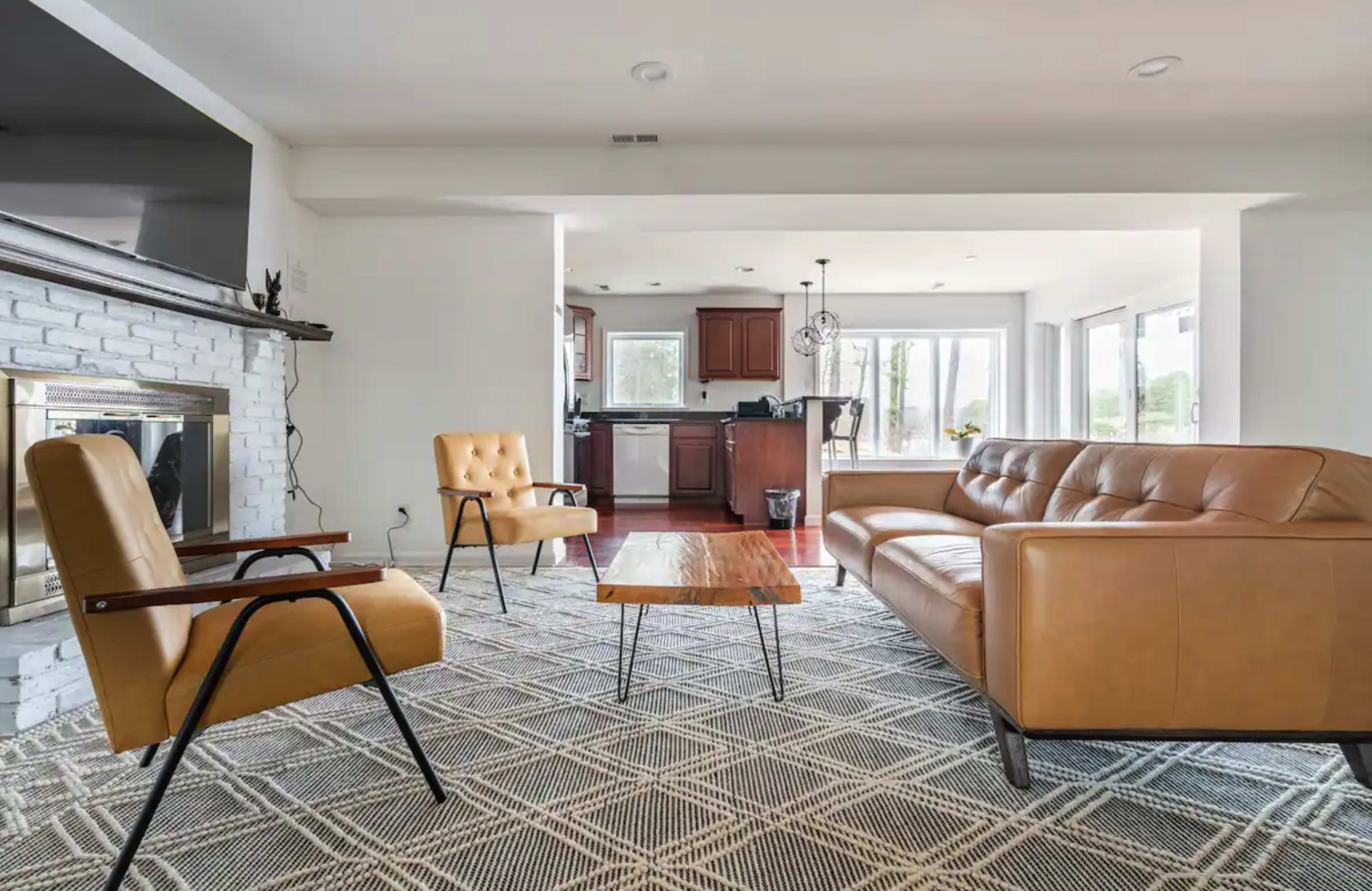 A modern living room with a tan leather couch, a wooden coffee table, and two armchairs, connected to a kitchen area with dark wood cabinets and large windows.