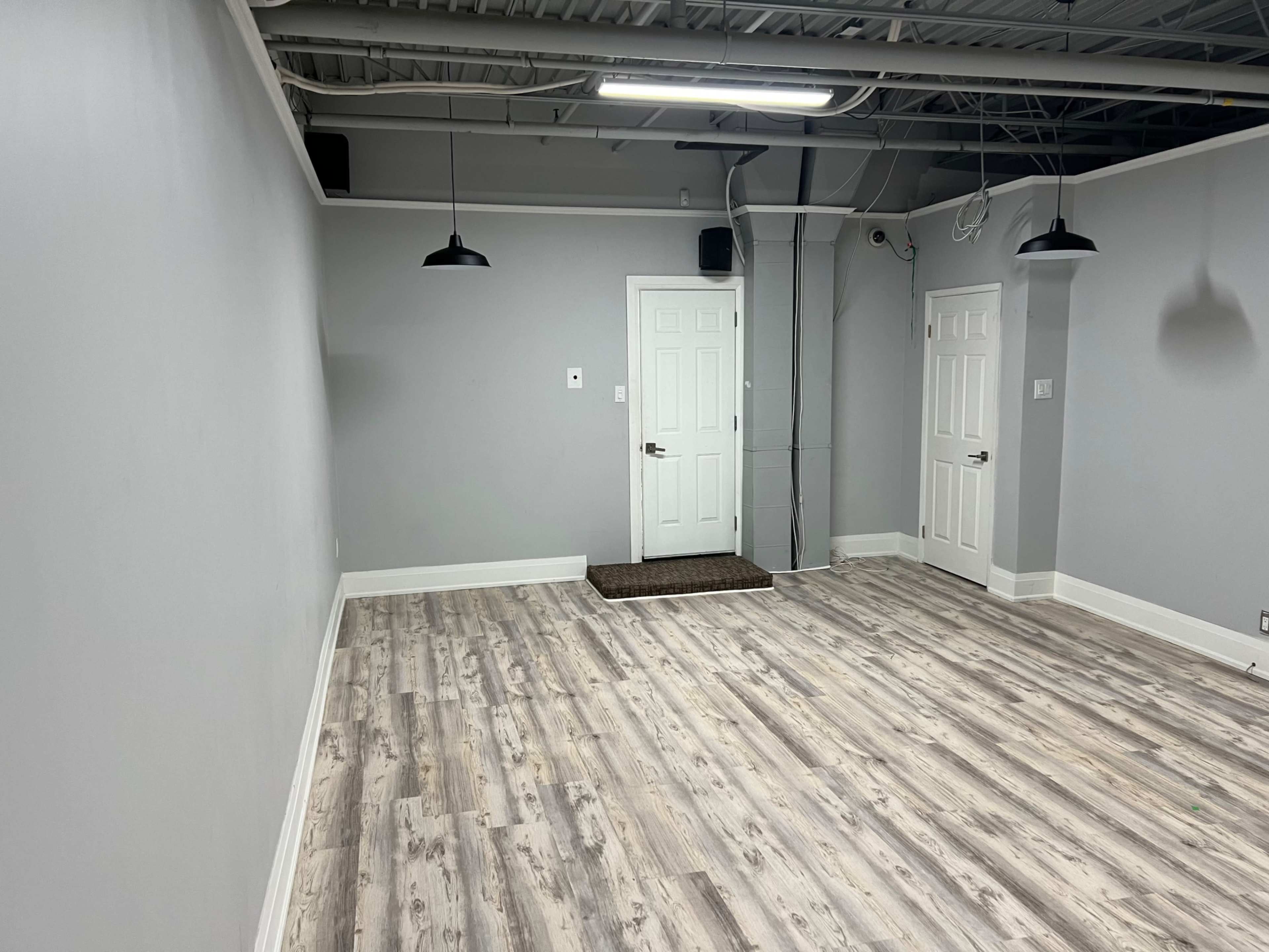 A vacant room with light gray walls, a wooden floor, and two doors, along with two pendant lights hanging from the ceiling.