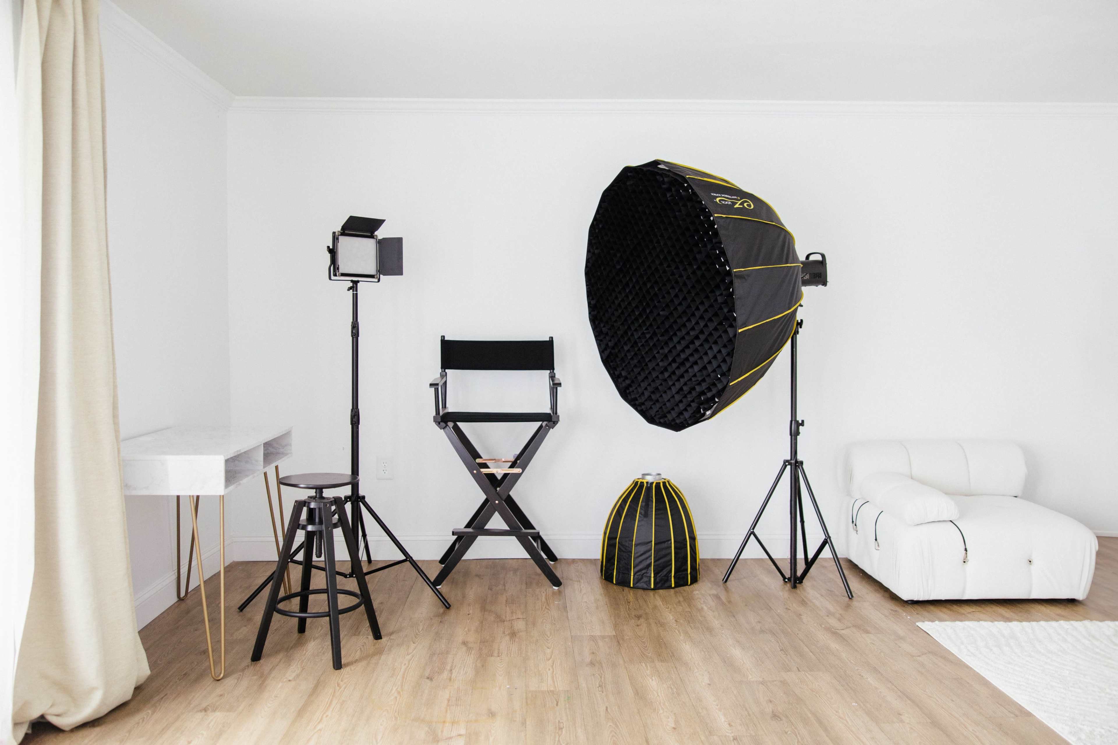 The scene shows a minimalist studio setting with a director's chair, lighting equipment, a large softbox, and a white sofa against a light-colored wall.