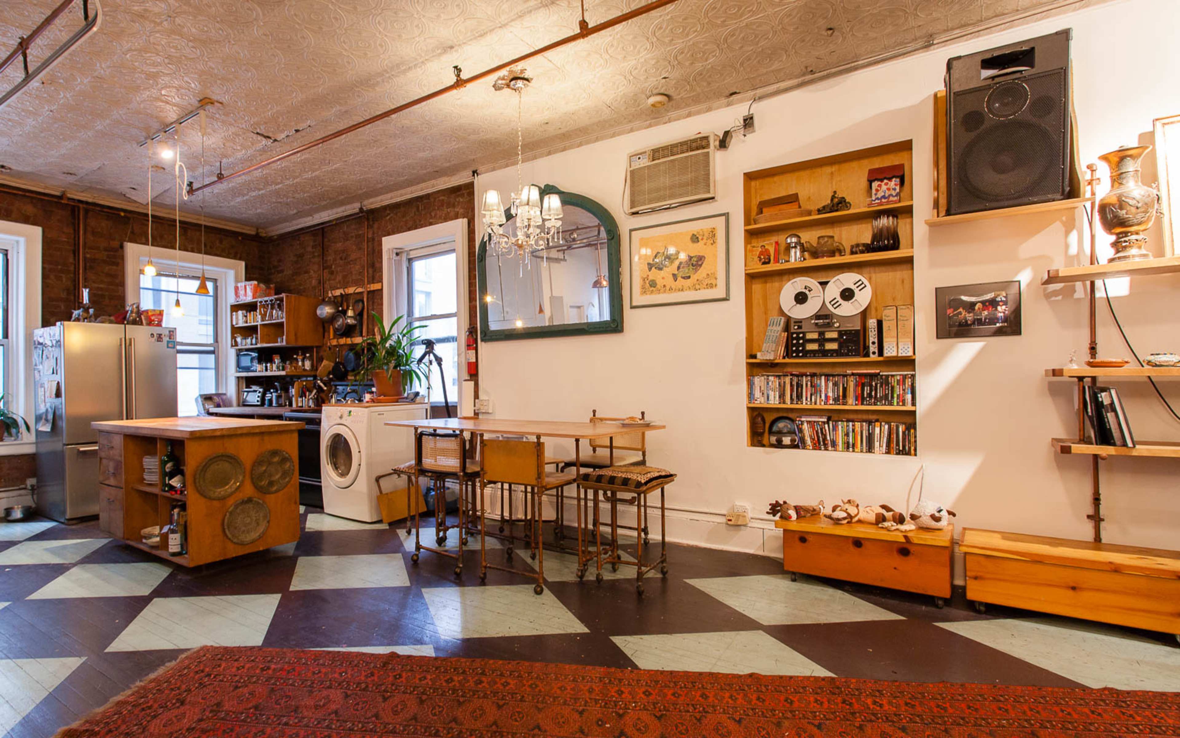 An open living space featuring a kitchen area with an island, a dining table, and shelves displaying various items, all set against a backdrop of patterned flooring.