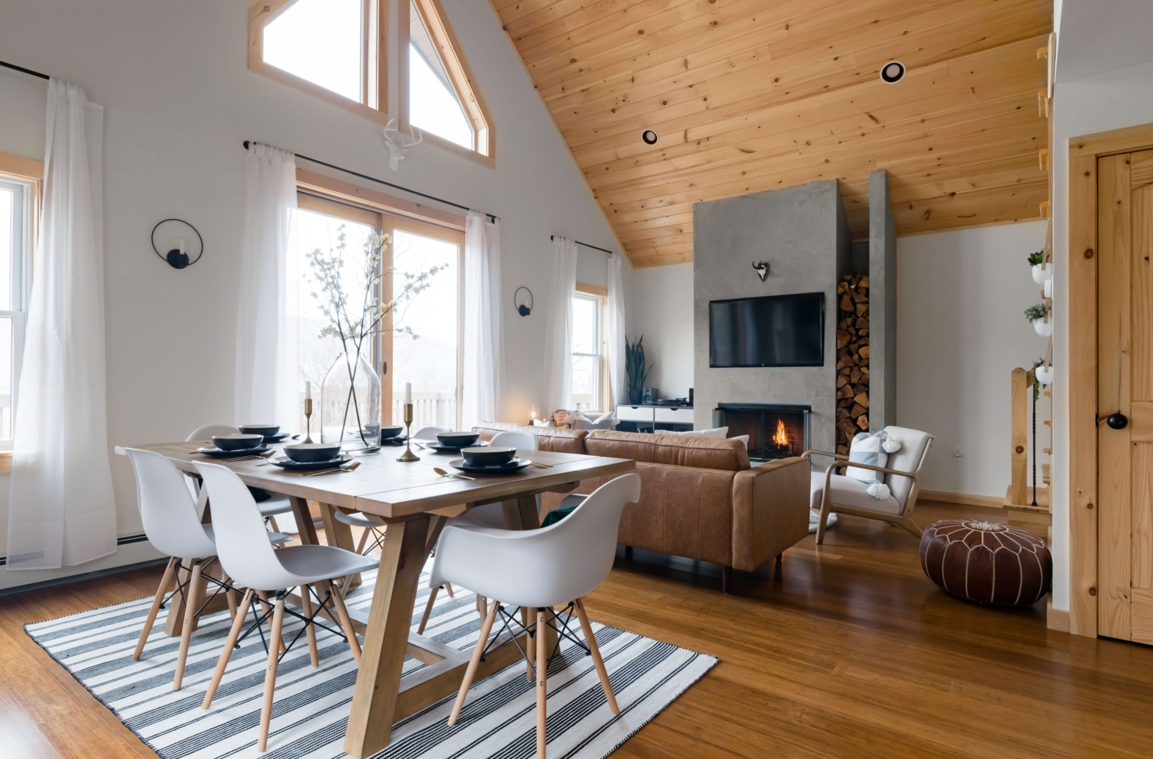 A modern living and dining area features a wooden table with stylish chairs, a cozy couch, and a fireplace under a high, vaulted ceiling with large windows.