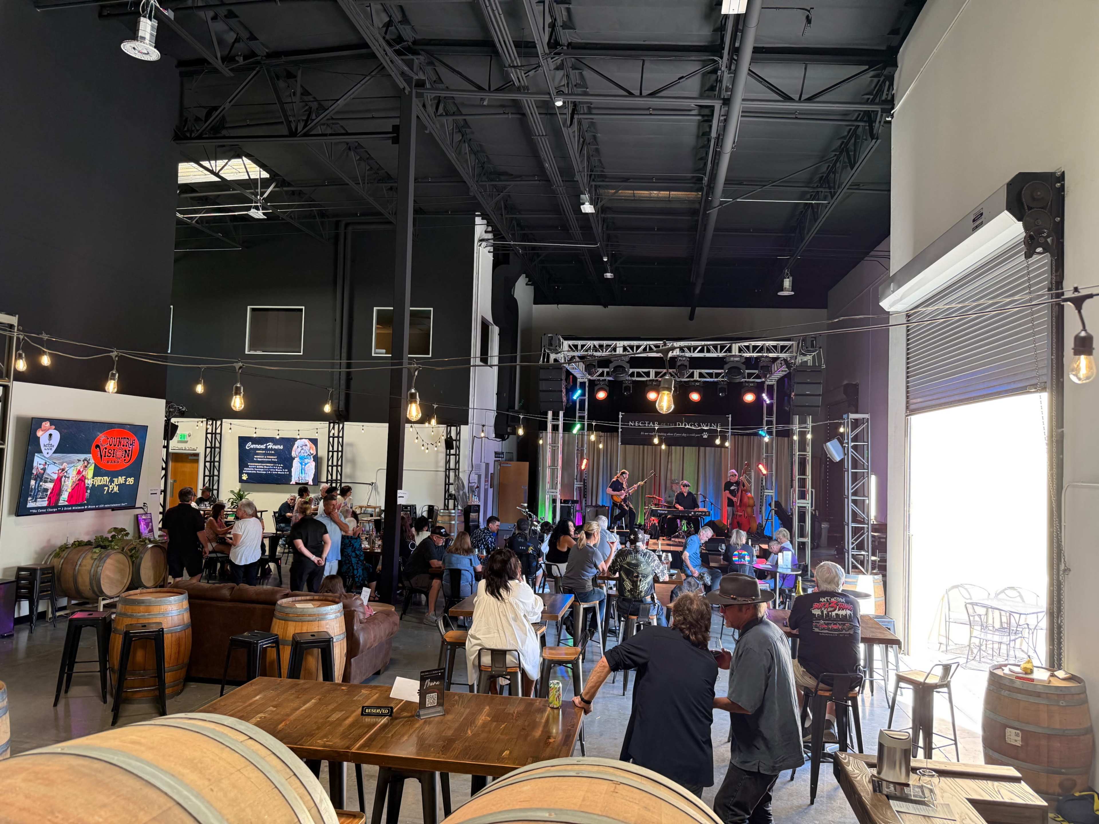 A crowd enjoys live music in a spacious venue with wooden barrels, tables, and a stage featuring performers.