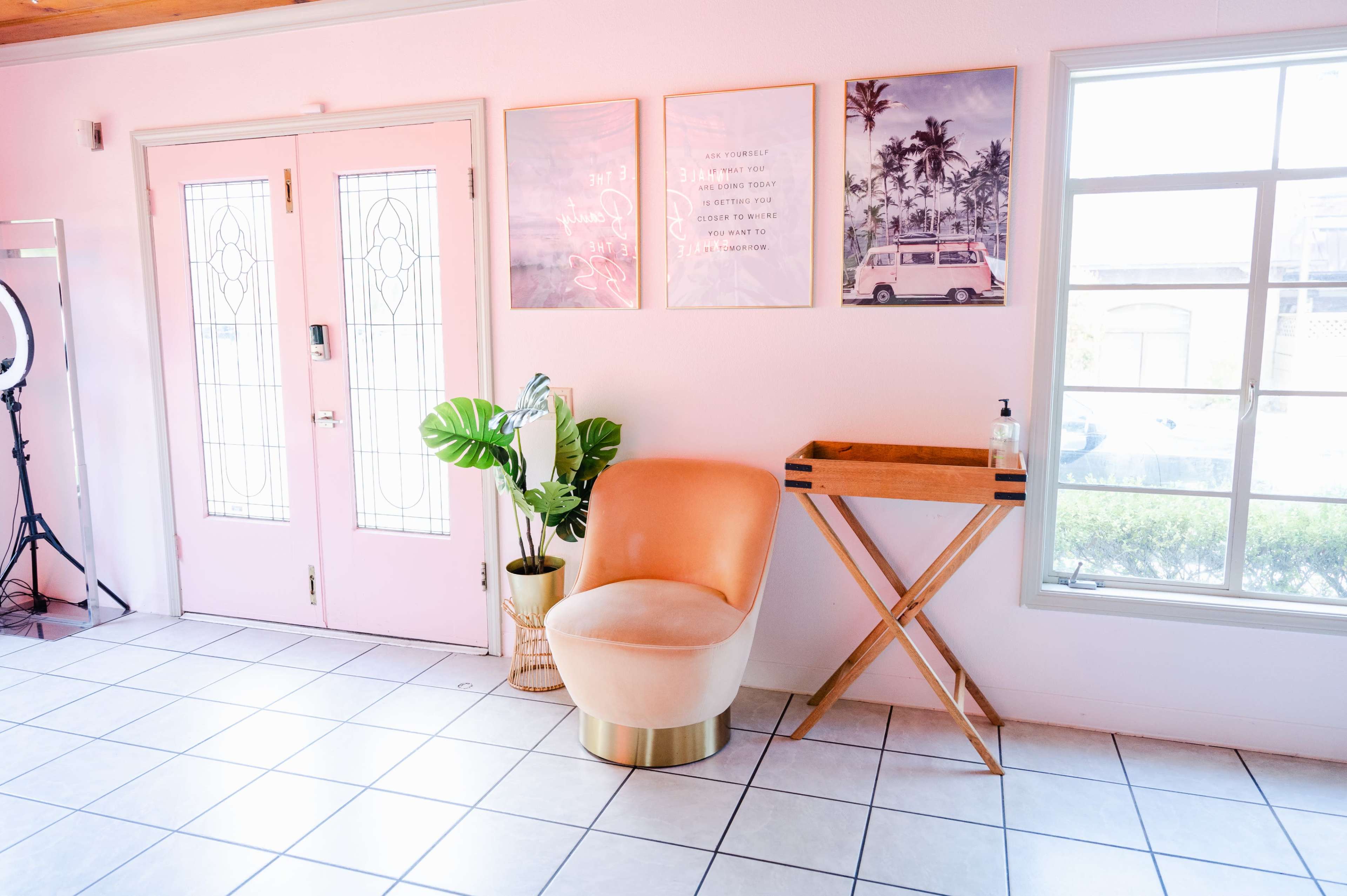 The image shows a bright interior space with a pink door, decorative wall art, a modern peach chair, and a wooden side table beside a large window.