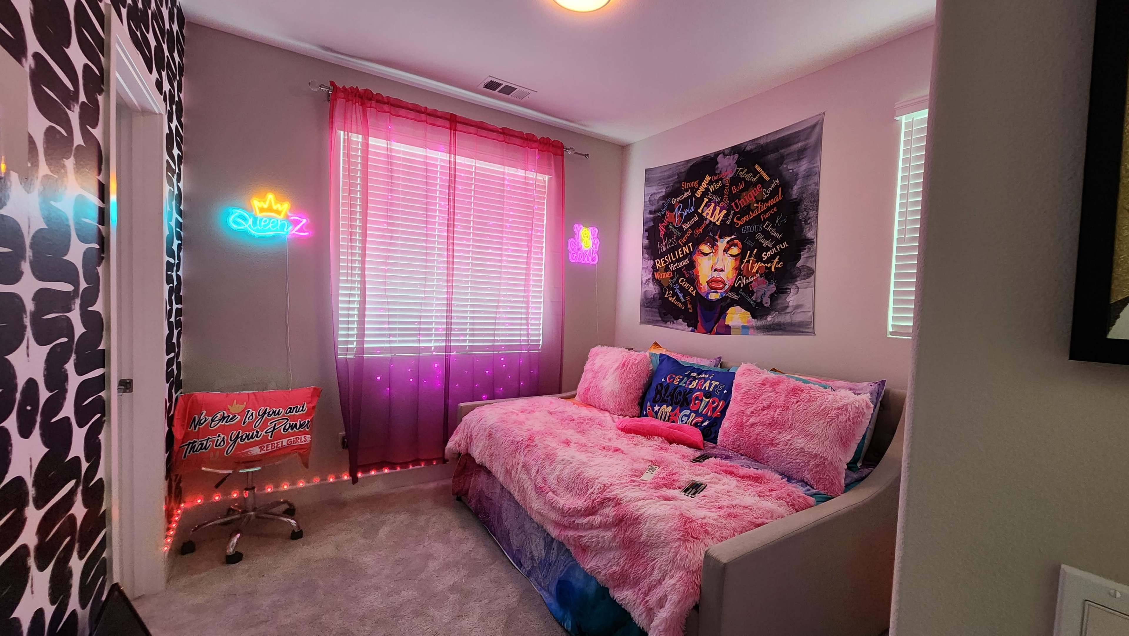 The room features a bed with a pink fur blanket, colorful wall art, and neon signs, along with large windows covered by sheer curtains.