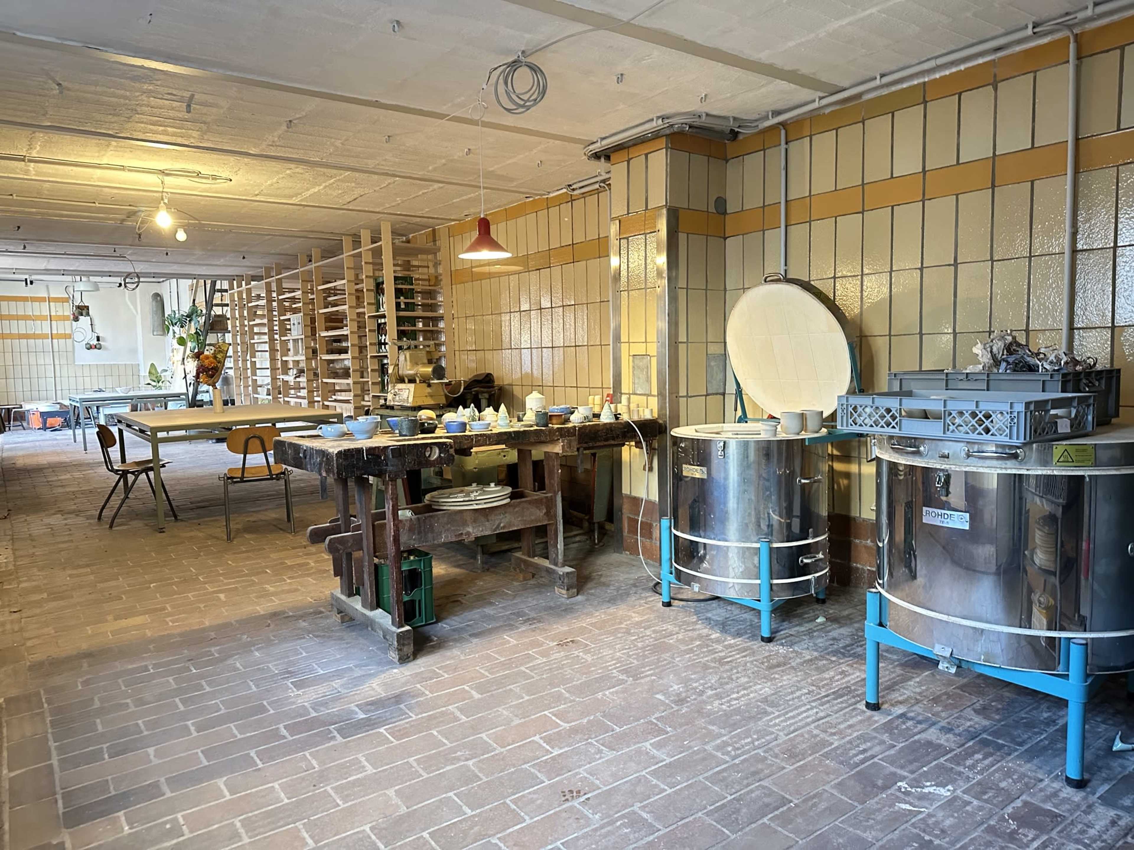 The image depicts a spacious workshop with various pottery tools and equipment, including large kilns and a worktable, under warm overhead lighting.