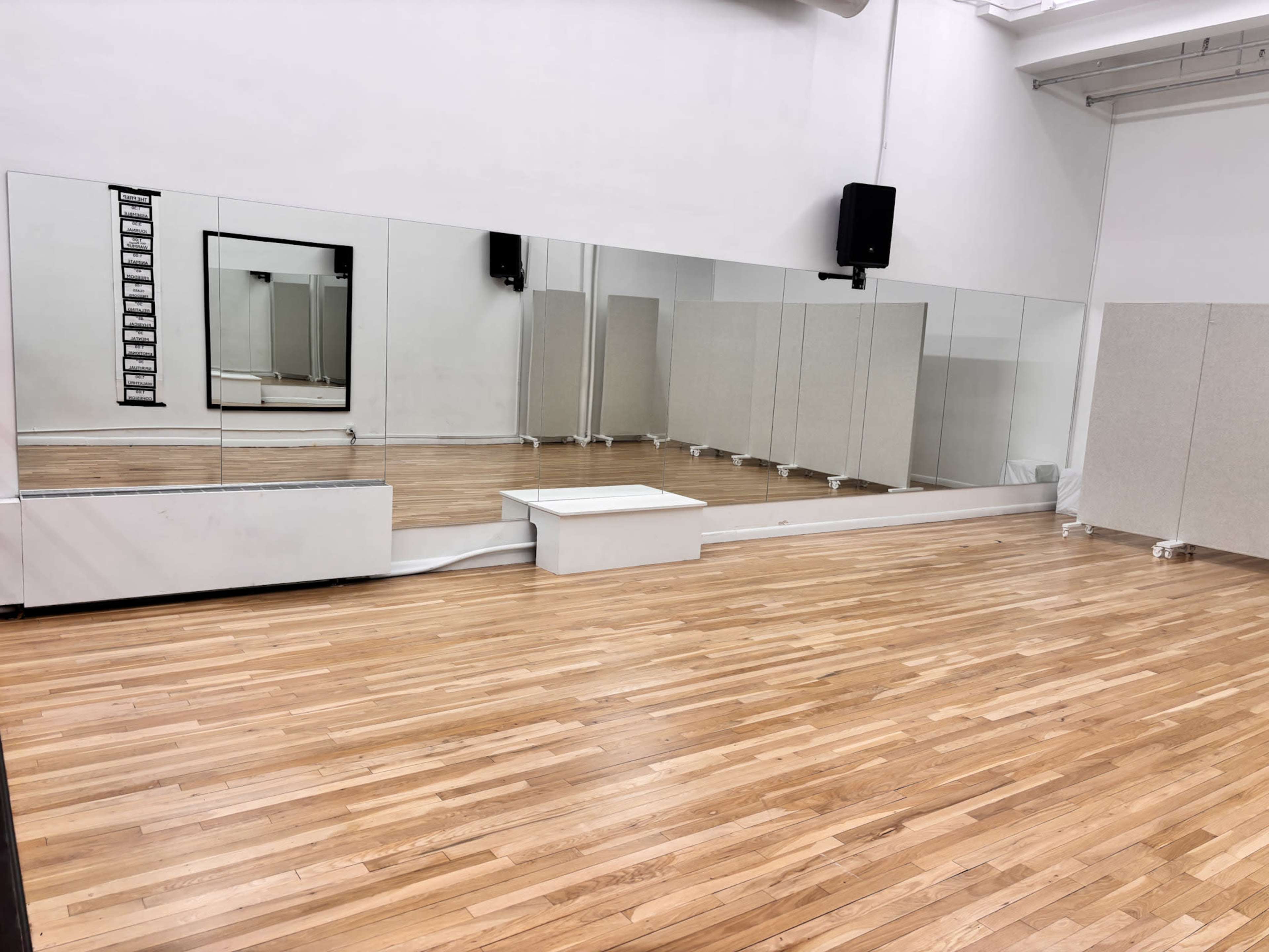 The image shows a spacious dance studio with wooden flooring, a large mirror on one wall, and sound equipment mounted in the corners.
