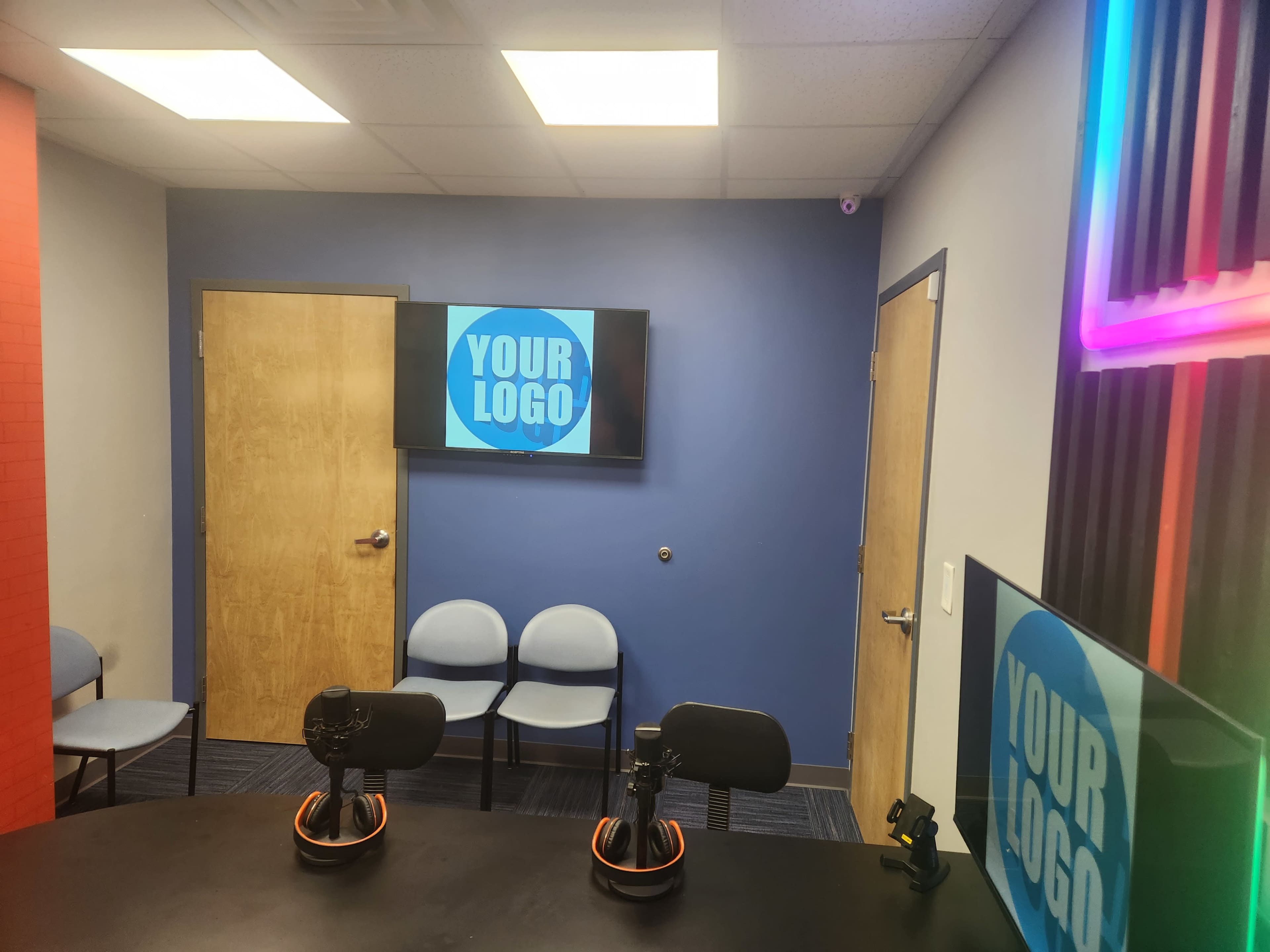 The image shows a studio or office space with two monitors displaying a logo, a table with microphones, and some chairs against the walls.