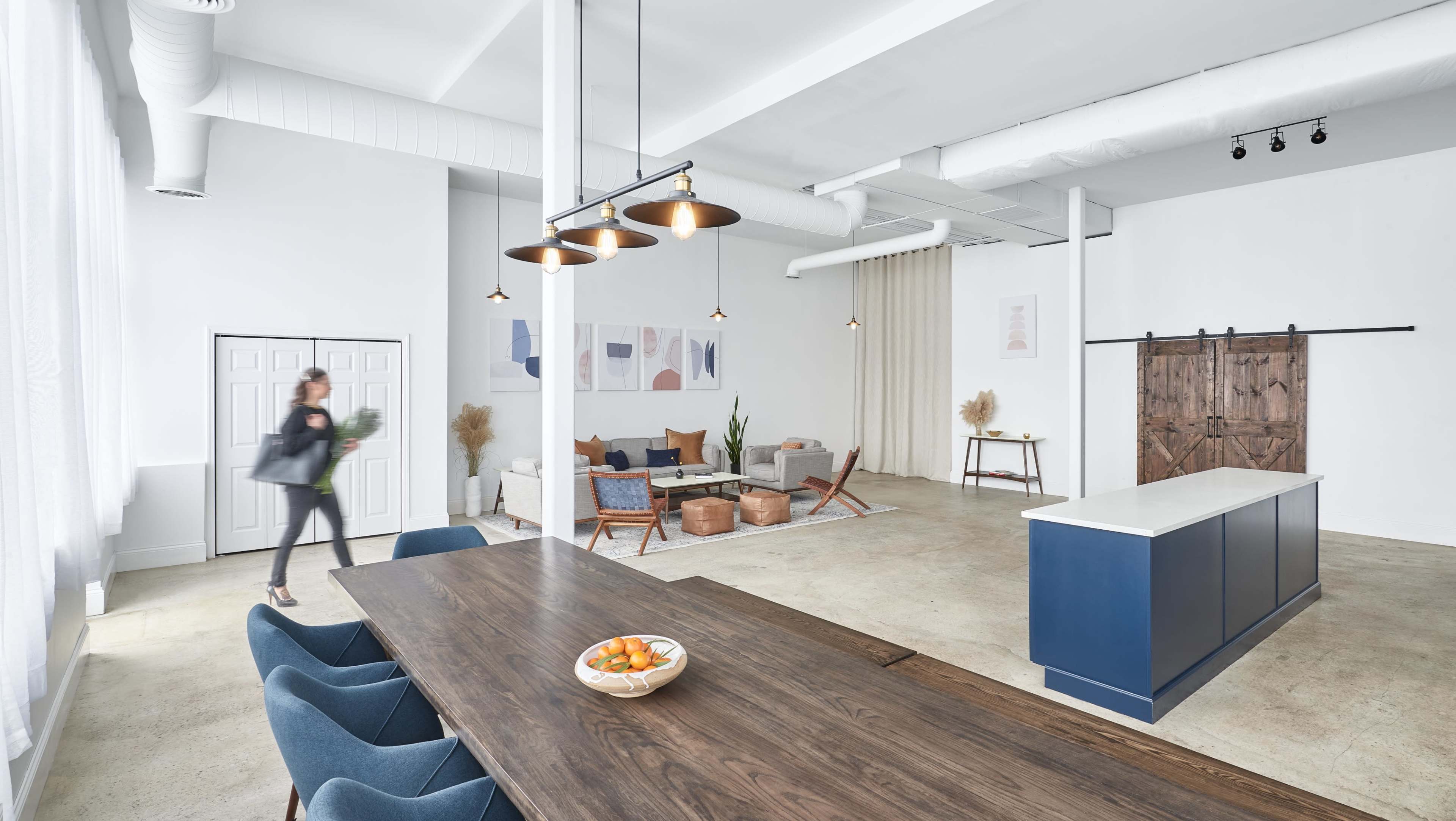 A spacious, modern living area features a dining table, a lounge with sofas, and a large door leading to another room, with a person walking through the space.