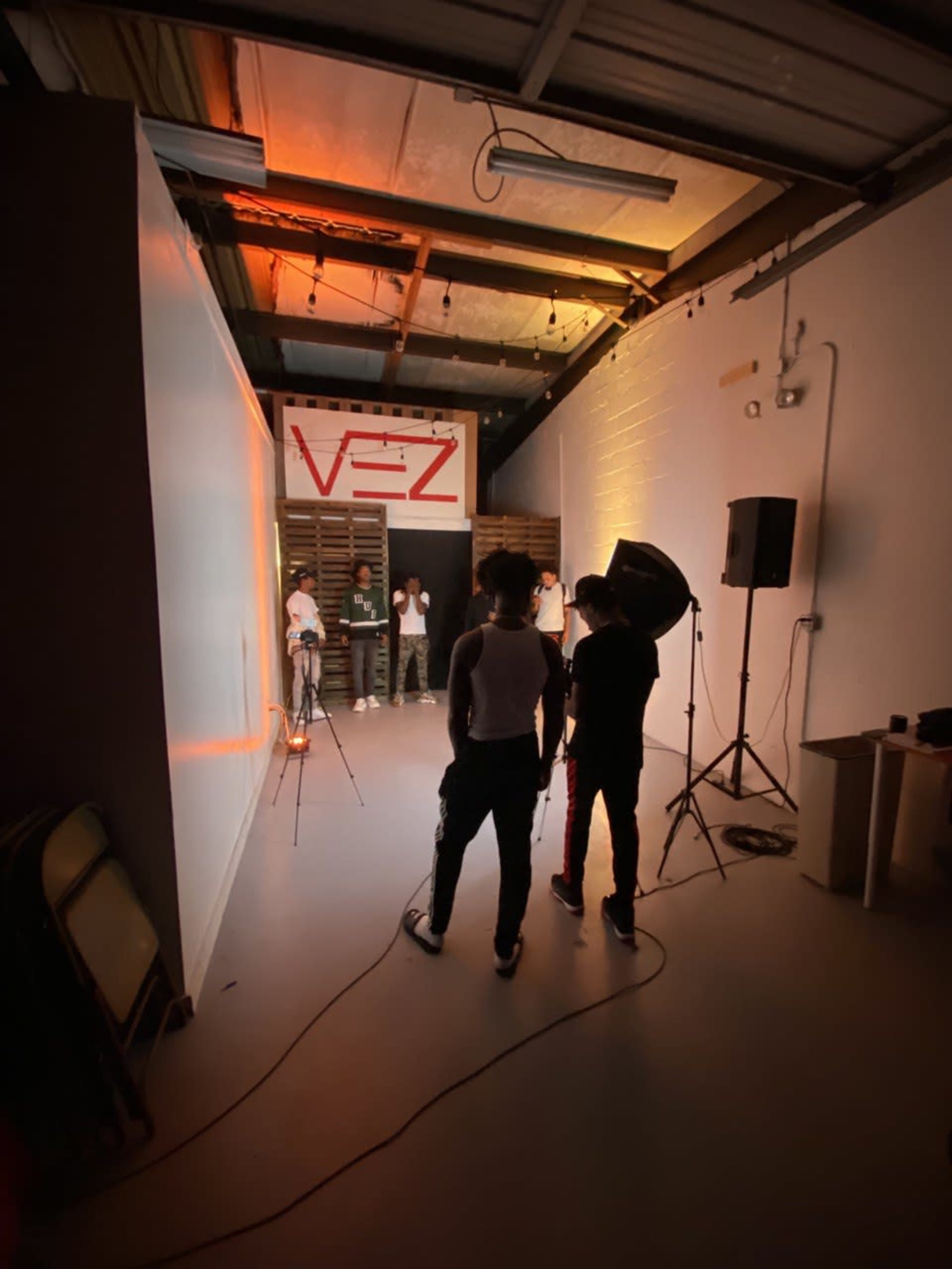 A photography studio features several individuals positioned in front of a backdrop displaying "V3Z," with equipment set up for a photoshoot.