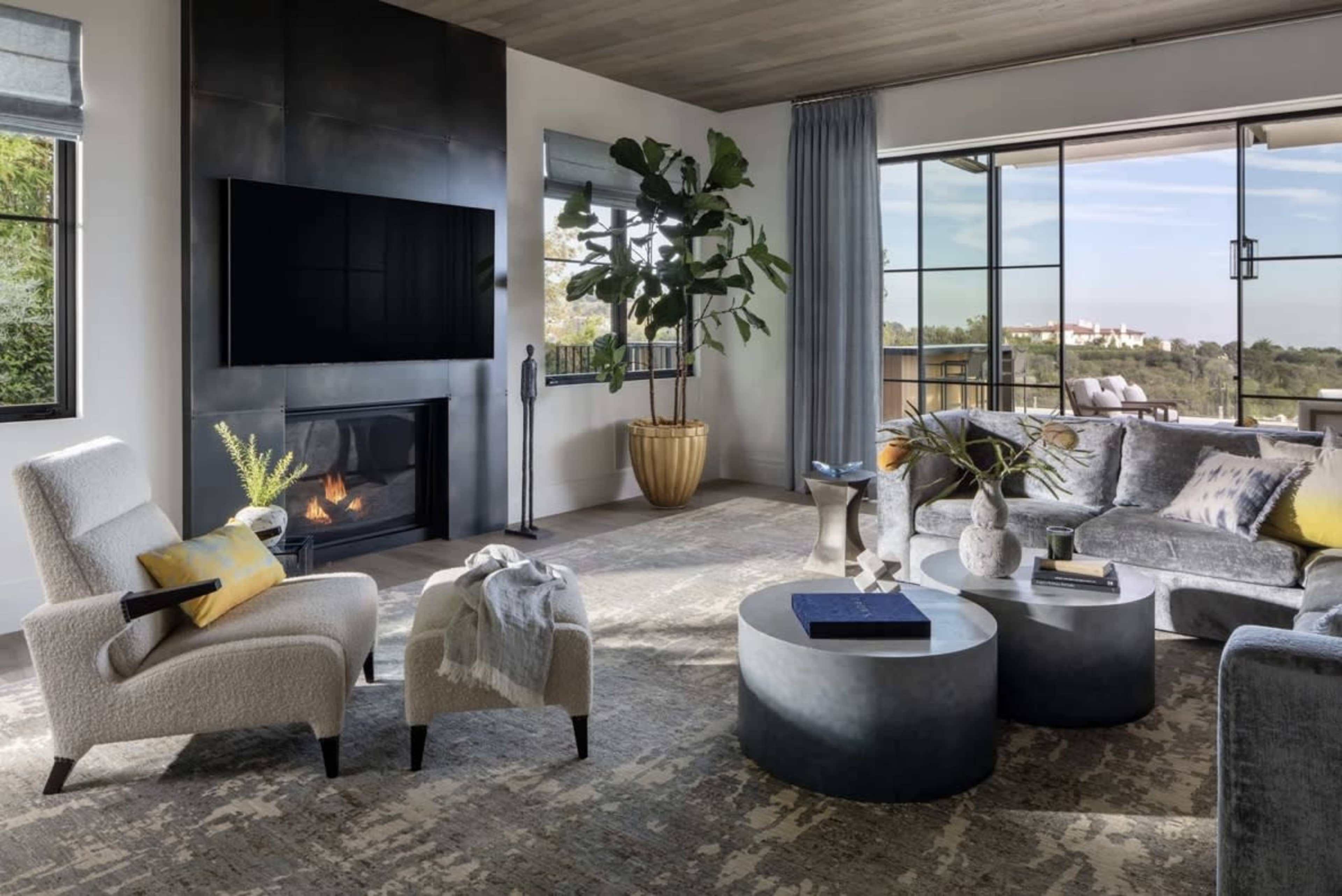 The image shows a modern living room featuring a fireplace, two armchairs, a sectional sofa, and large windows that overlook a scenic view.